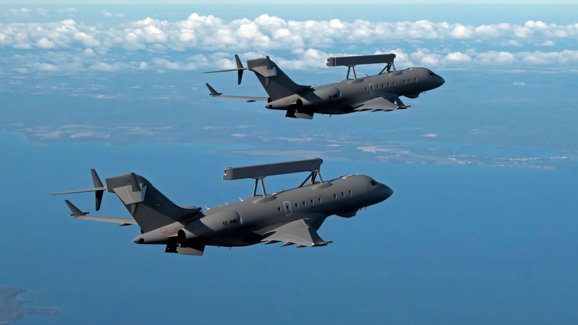 A pair of GlobalEye aircraft fly over water and specks of land. White clouds are seen in the background.