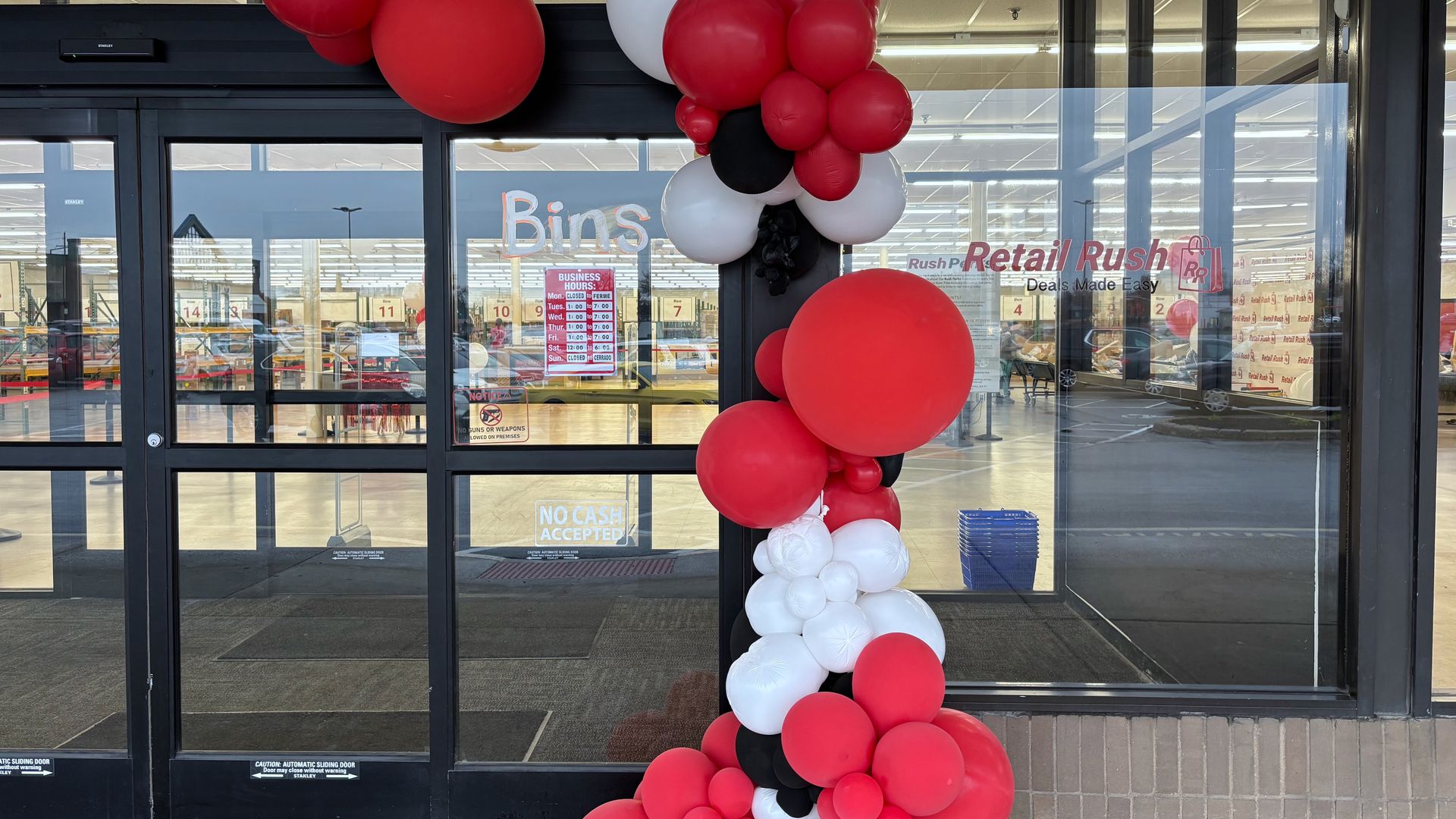 Store entrance decorated with a balloon column of red, white, and black balloons; glass doors with business hours and signs, inside a retail store visible.