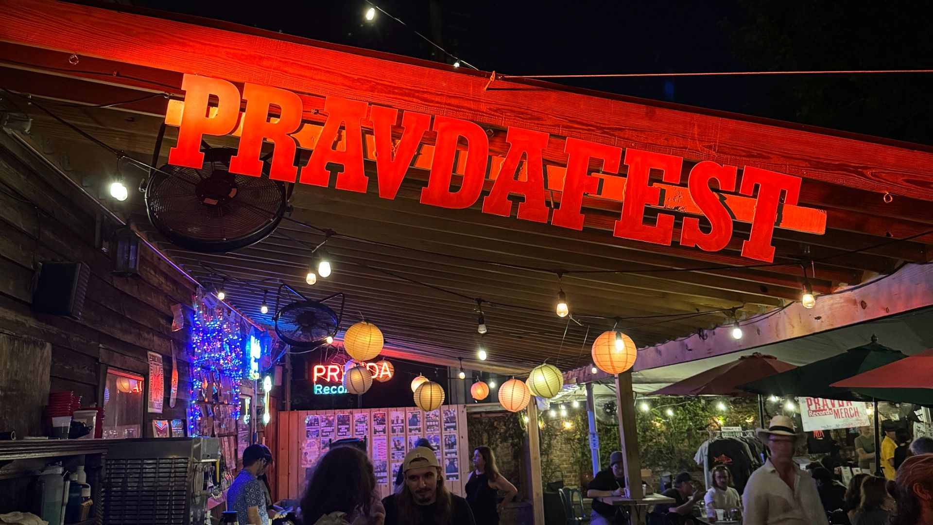 Night market at night with a large red sign reading "PRAVDAFEST" across a wooden beam; string lights, hanging lanterns, ceiling fans, and people dining outdoors.