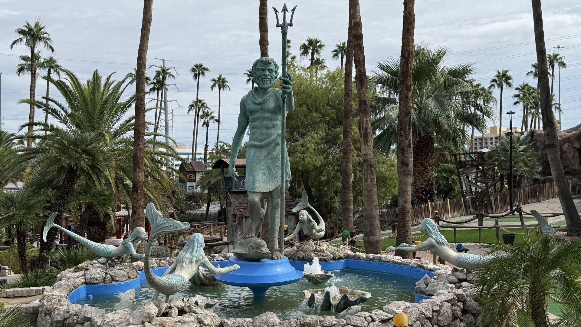 Green statue of Poseidon with trident surrounded by four mermaid sculptures in a blue water fountain, with palm trees in the background.
