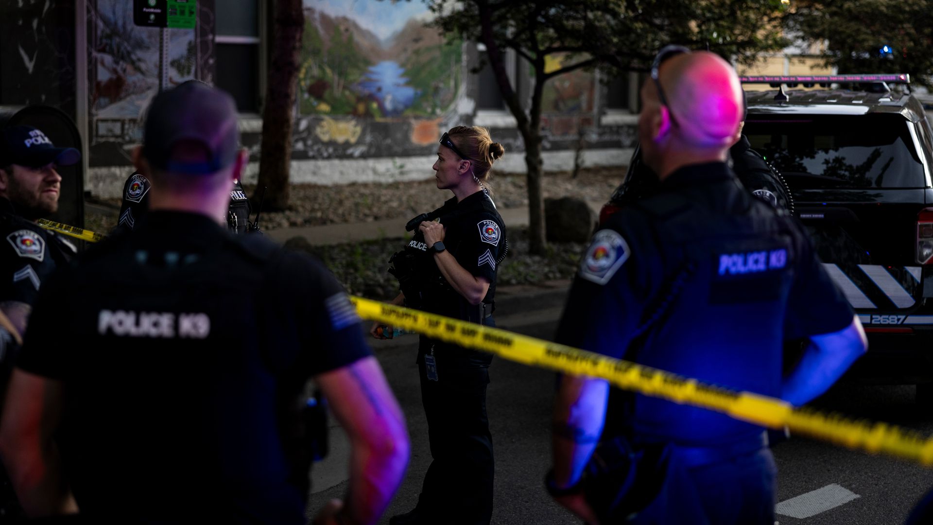 Two police officers in the foreground and another in the background, standing around police cards, yellow police tape and trees.