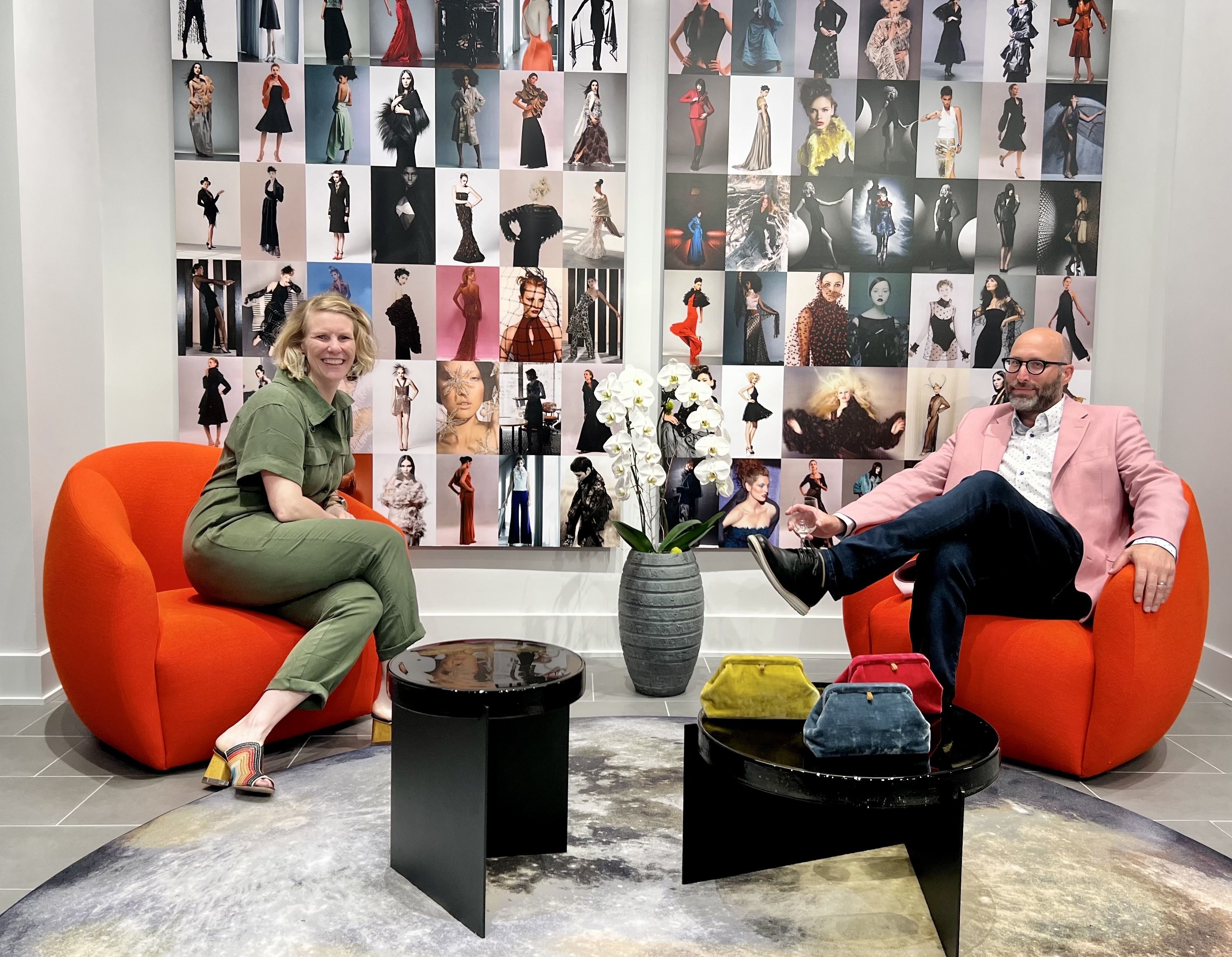 Wall with collage of models behind woman in green jumpsuit siting in orange chair next to man in pink blazer sitting in orange chair. Table in center with vase and flowers.