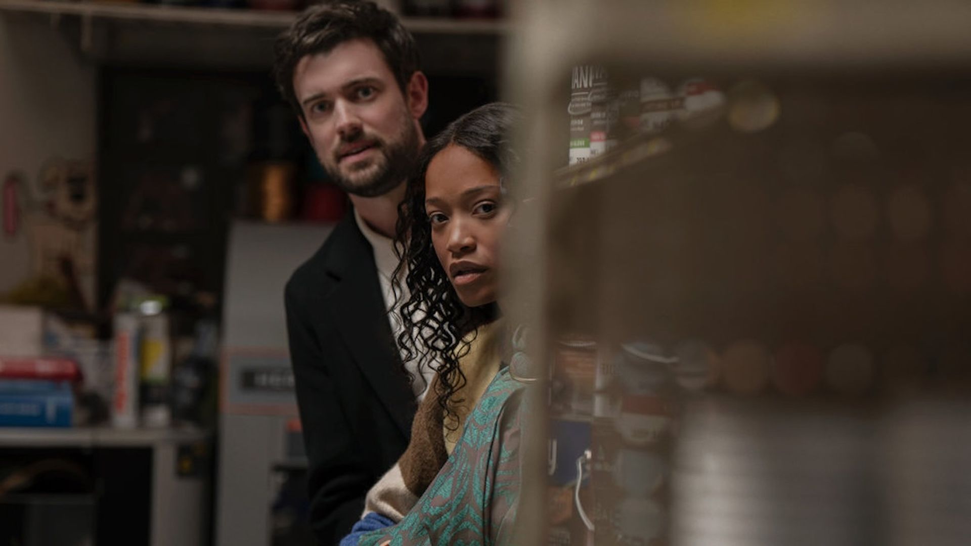A man with dark hair and beard wearing a black jacket stands behind a woman in a patterned shawl, both looking intently forward inside a dimly lit room with shelves on the right.