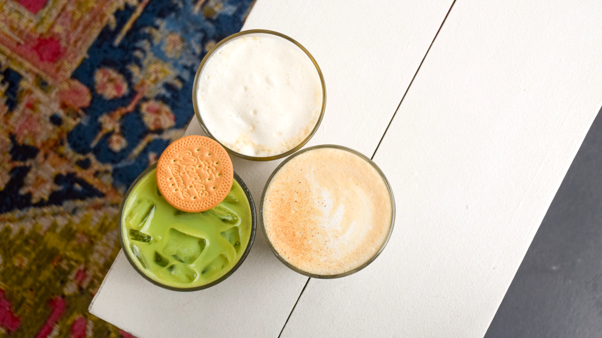 Top view of three drinks on a white wooden table: a green iced matcha latte with a beige biscuit on top, a frothy white coffee, and a creamy beige latte with foam art, next to a colorful patterned rug.