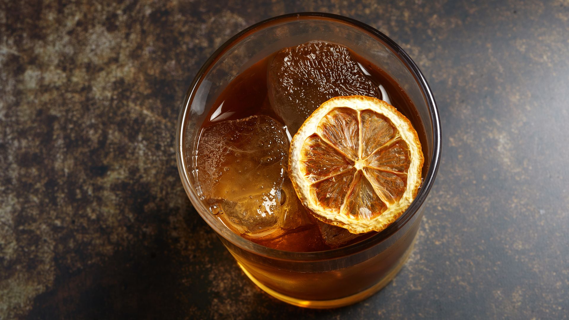 A lowball glass sits on a marbled counter top. It is viewed from above and the glass is filled with an orange liquid, a dried orange slice and ice.