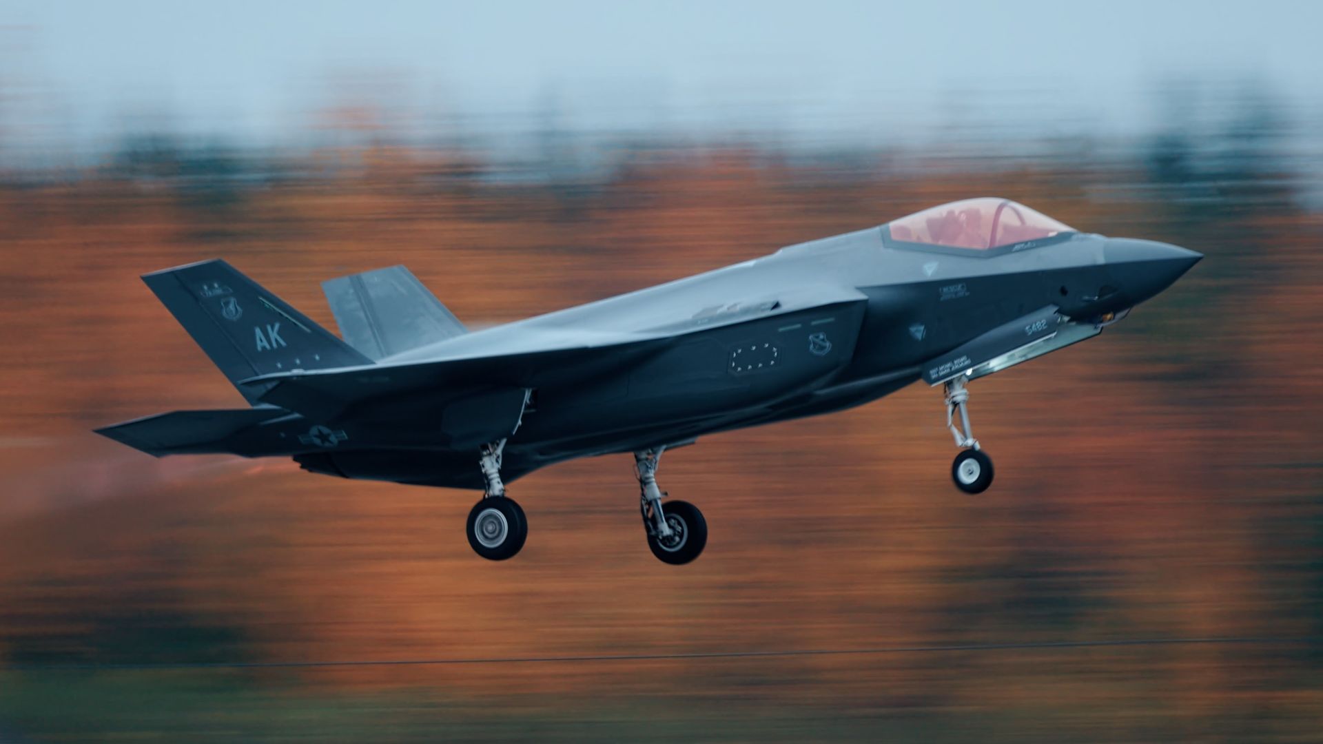 An F-35 takes off at a high speed. The background, a dreary fall scene, is blurred.