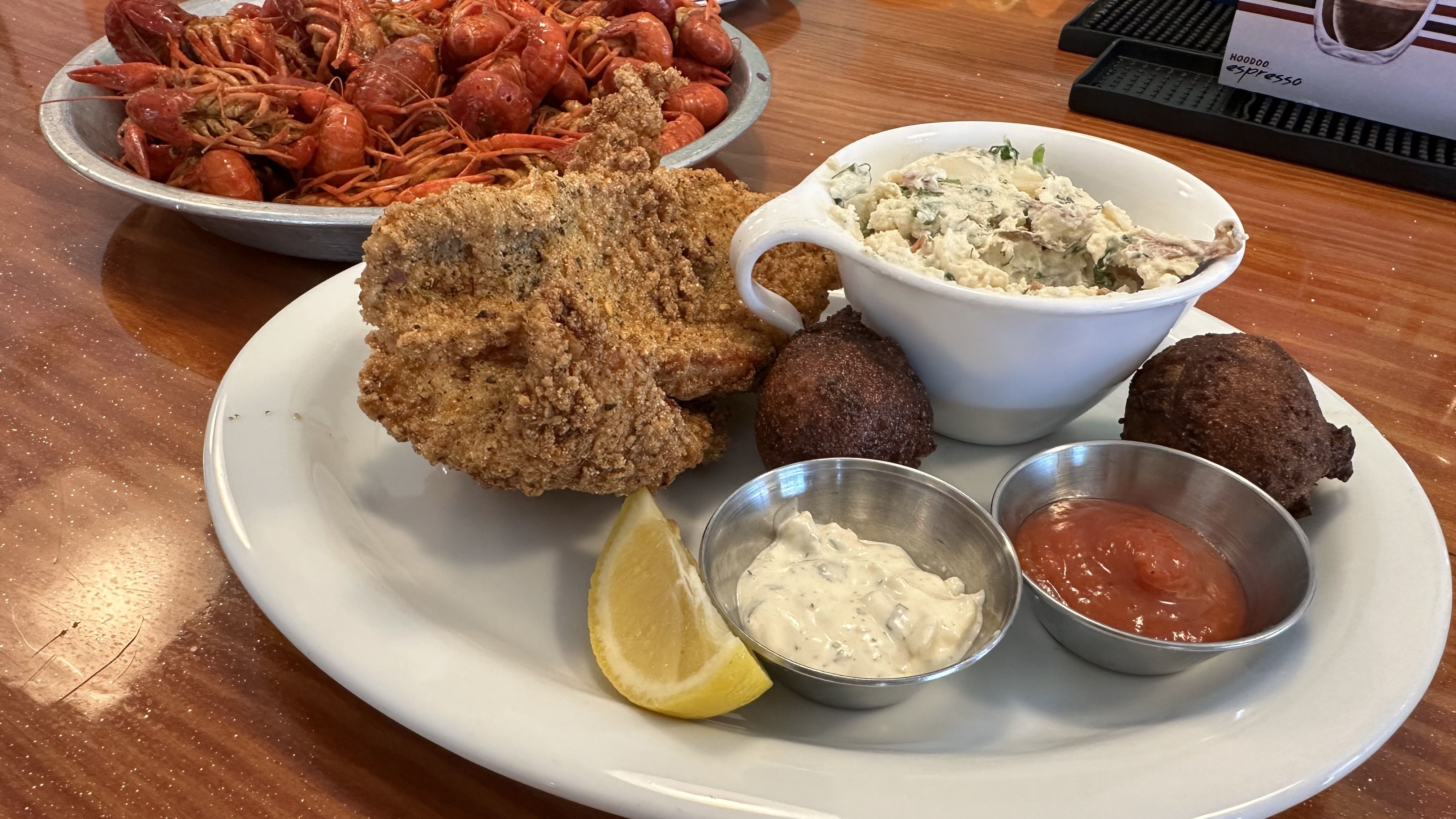 A white plate sits on a wooden bar. The plate is filled with fried catfish, a dish of potato salad, two hush puppies, a lemon wedge, and small silver containers of tartar sauce and cocktail sauce. Behind the plate is a large dish of boiled crawfish.