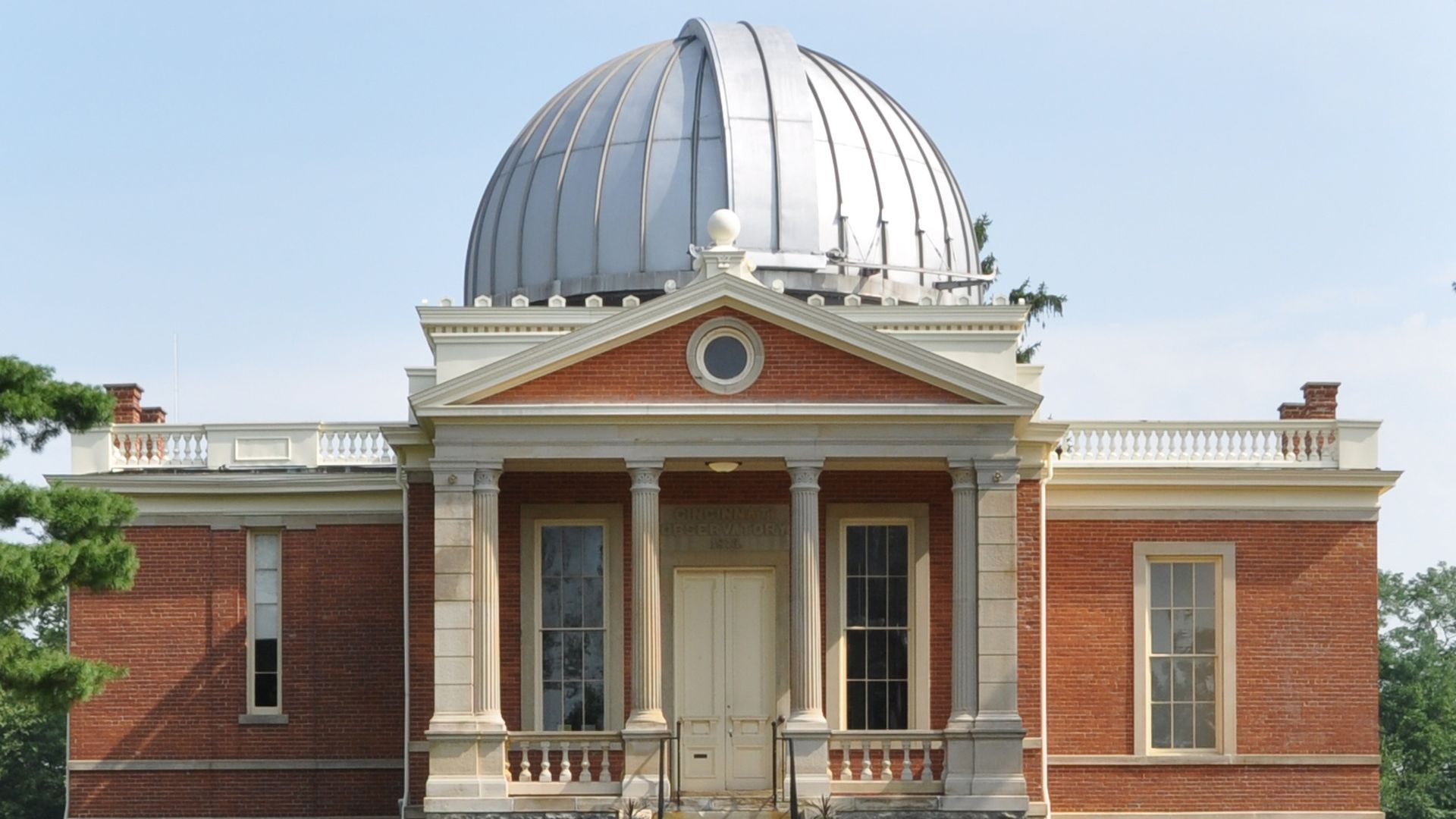 Red brick neoclassical building with white trim and a large silver dome. Front entrance features four columns and a triangular pediment, with a circular window and stone foundation on a green lawn.