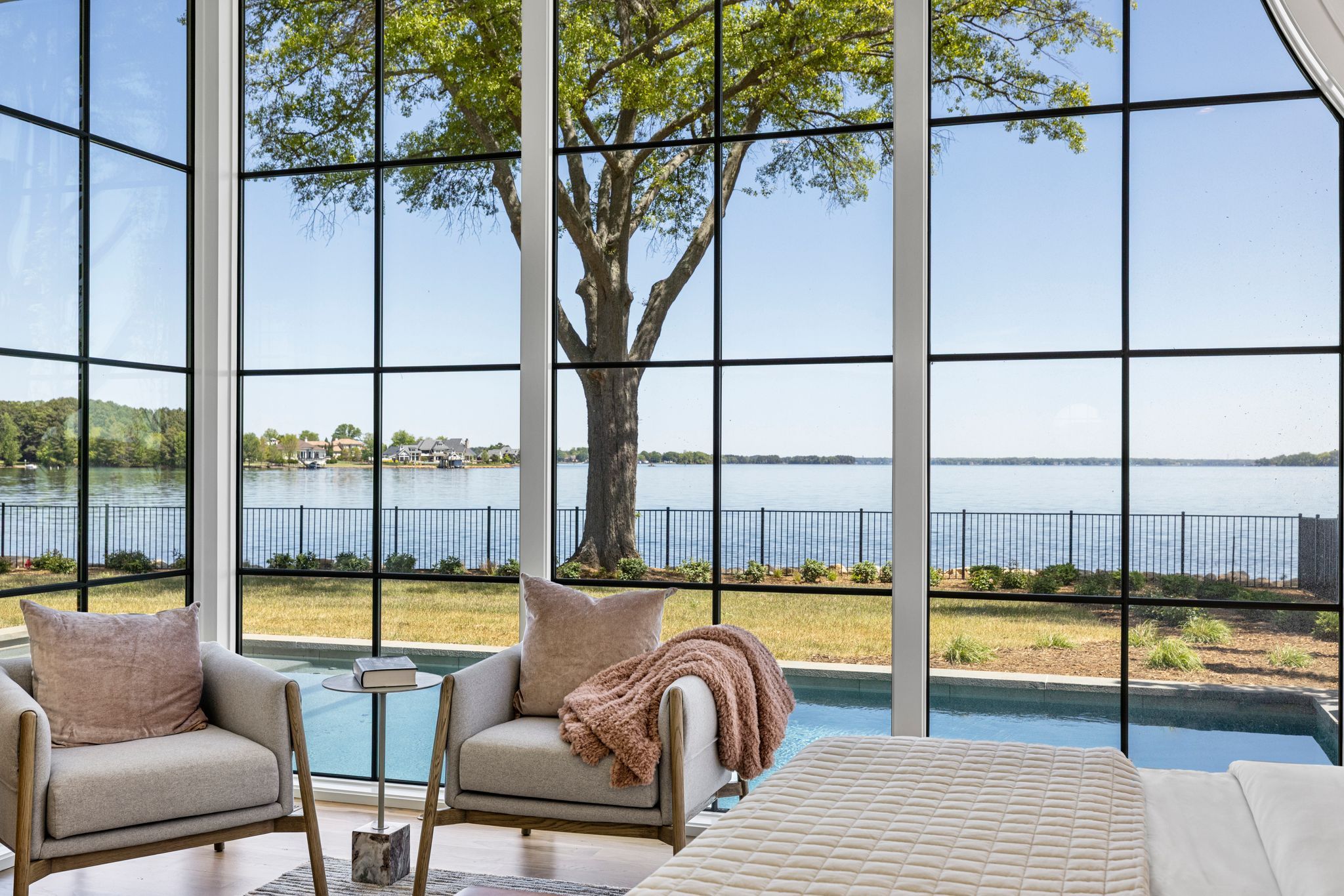 Cozy seating area with two gray armchairs and a small table, looking out through large black-framed windows at a tree, a grassy yard, a pool, and a calm blue lake under a clear sky.