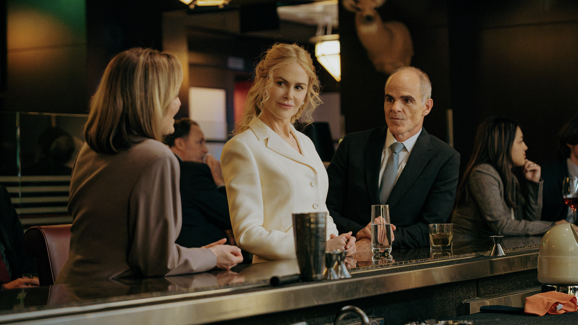 Three people in formal attire talk at a dimly lit bar with a mounted deer head above. The woman in white faces a man in a dark suit holding a glass of water.