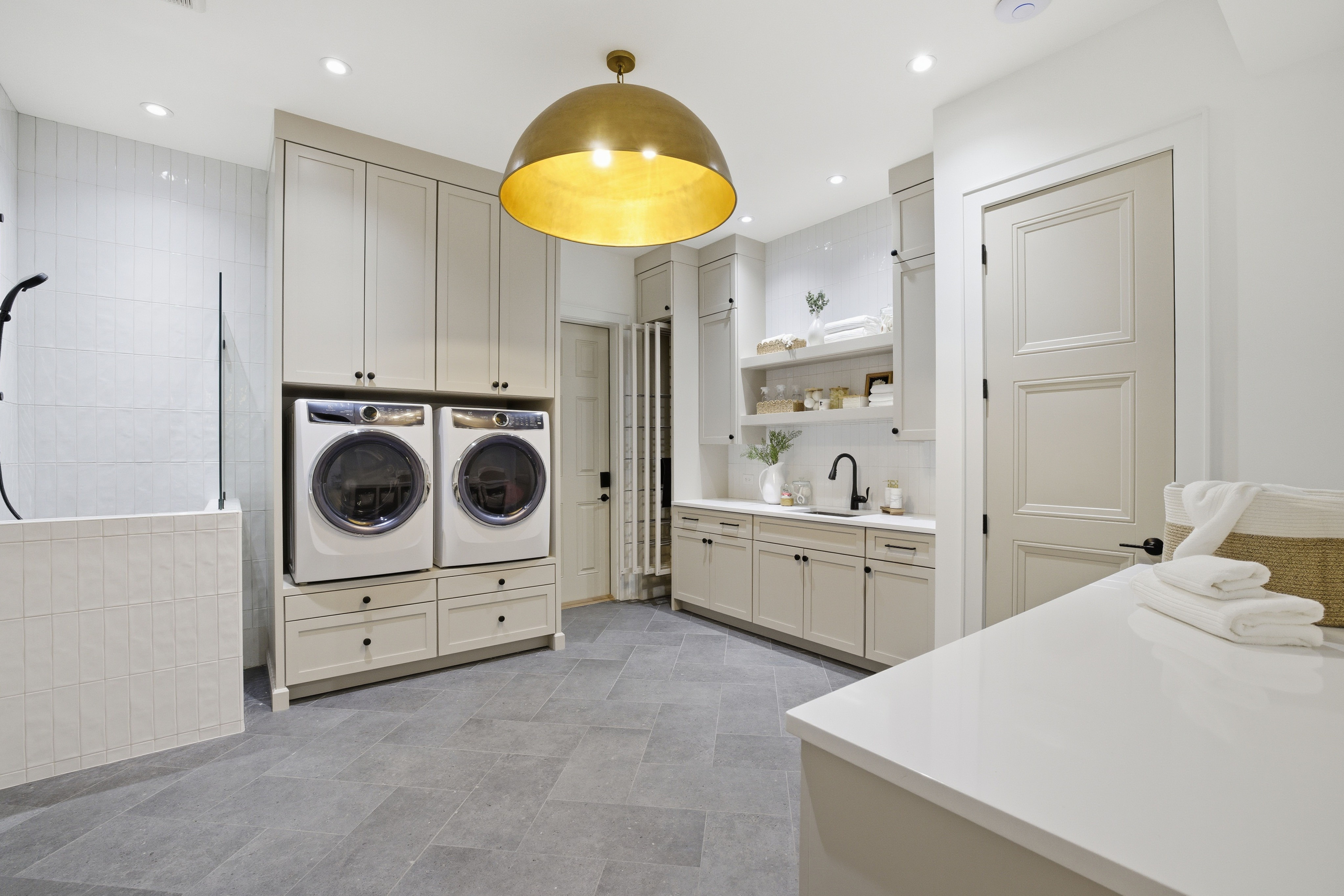Modern laundry room with gray cabinets, front-loading washer and dryer, white countertops, black faucet, open shelves with towels and decor, gray tile floor, and a large gold pendant light.