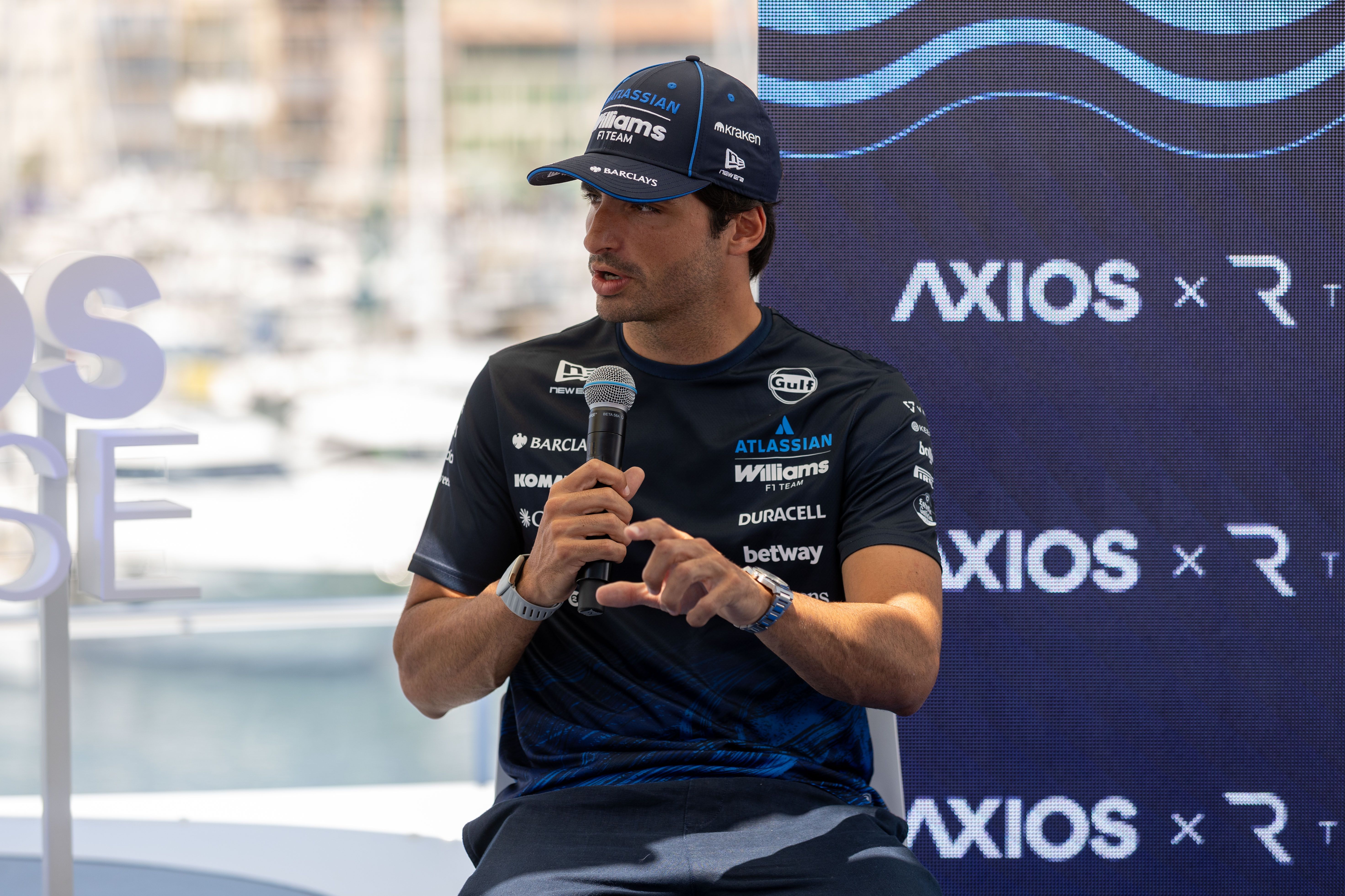 A man wearing a dark blue Williams F1 Team shirt and cap sits and speaks into a microphone during a press event, with a blue wave-pattern backdrop showing the word "AXIOS" behind him.