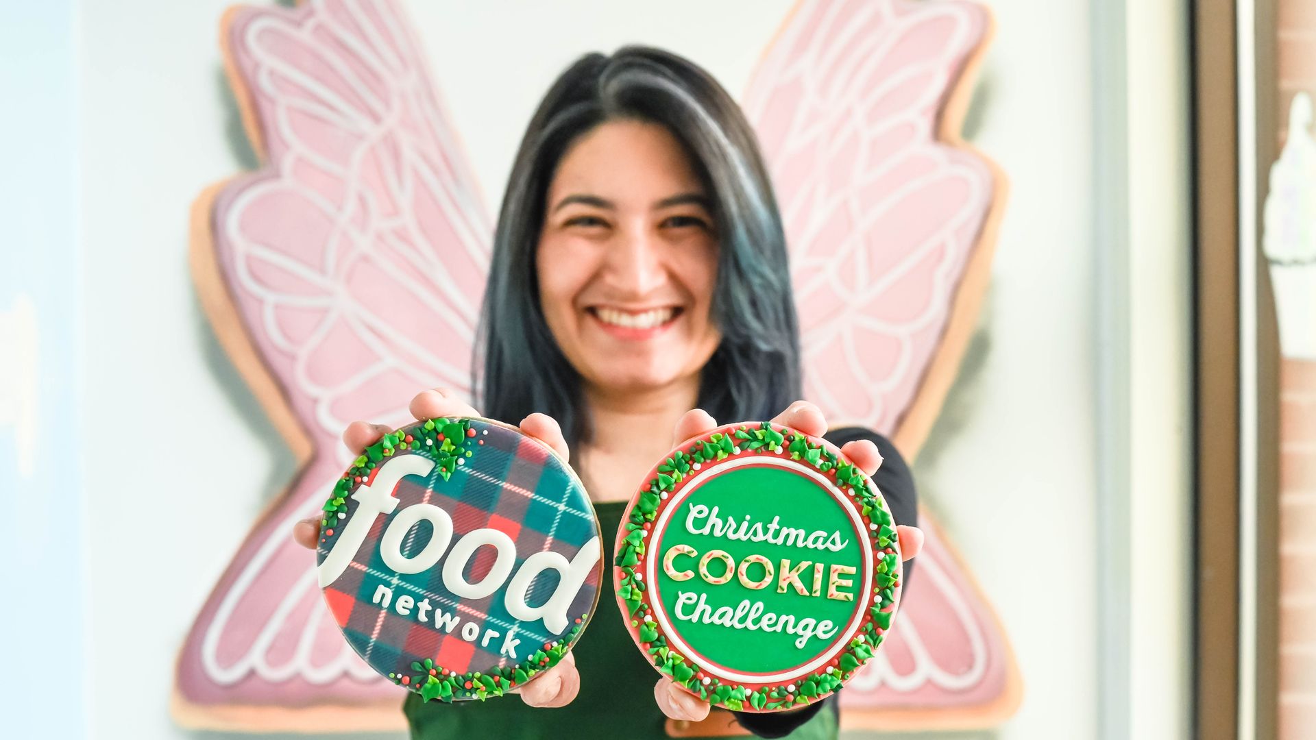 Smiling woman with dark hair holds two round decorated cookies: one with "food network" on red and green plaid, the other green with "Christmas COOKIE Challenge", with pink butterfly wings background.