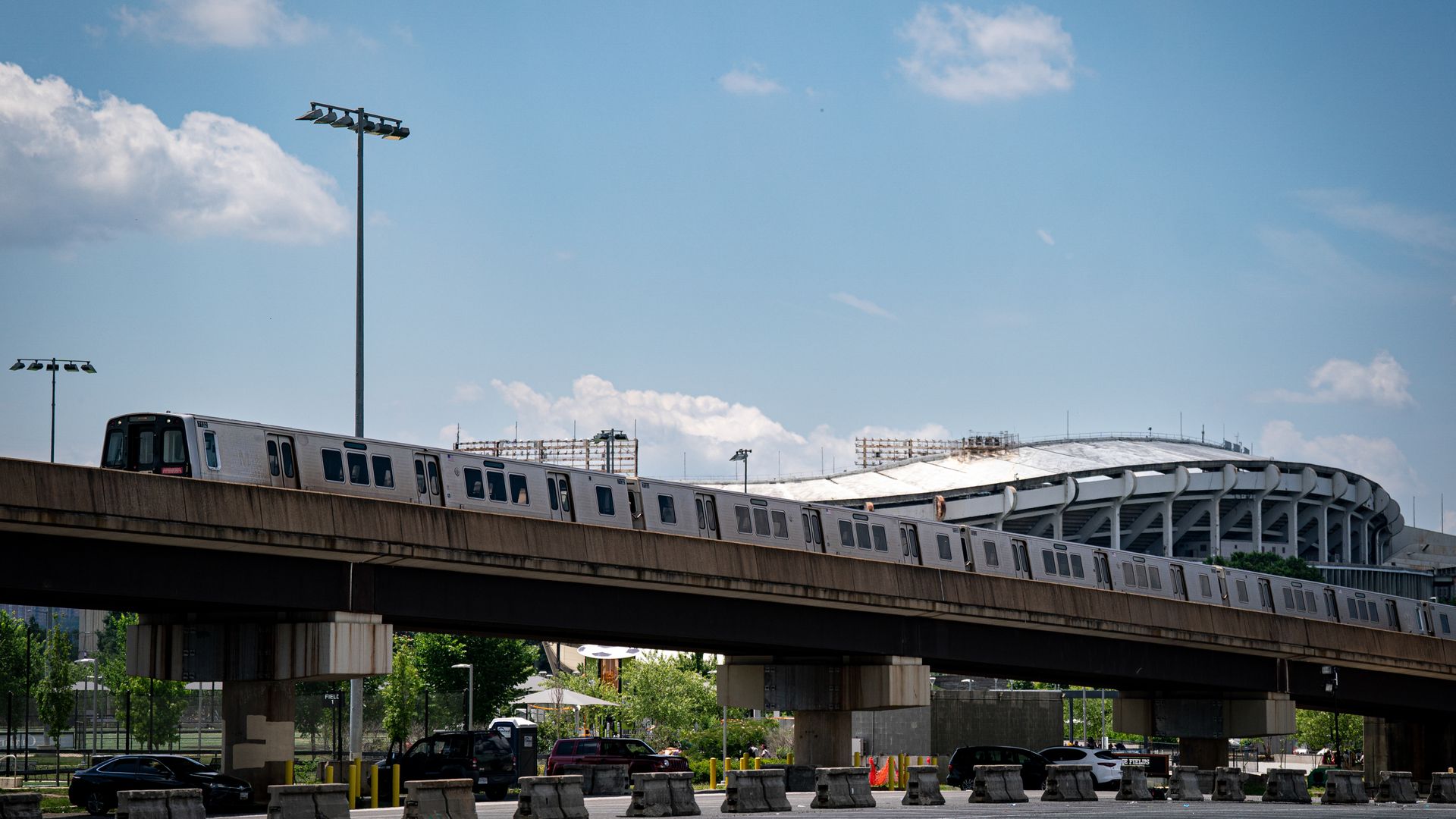 Silver subway train crosses an elevated track over a street with concrete barriers, with RFK Stadium and bright blue sky in the background.