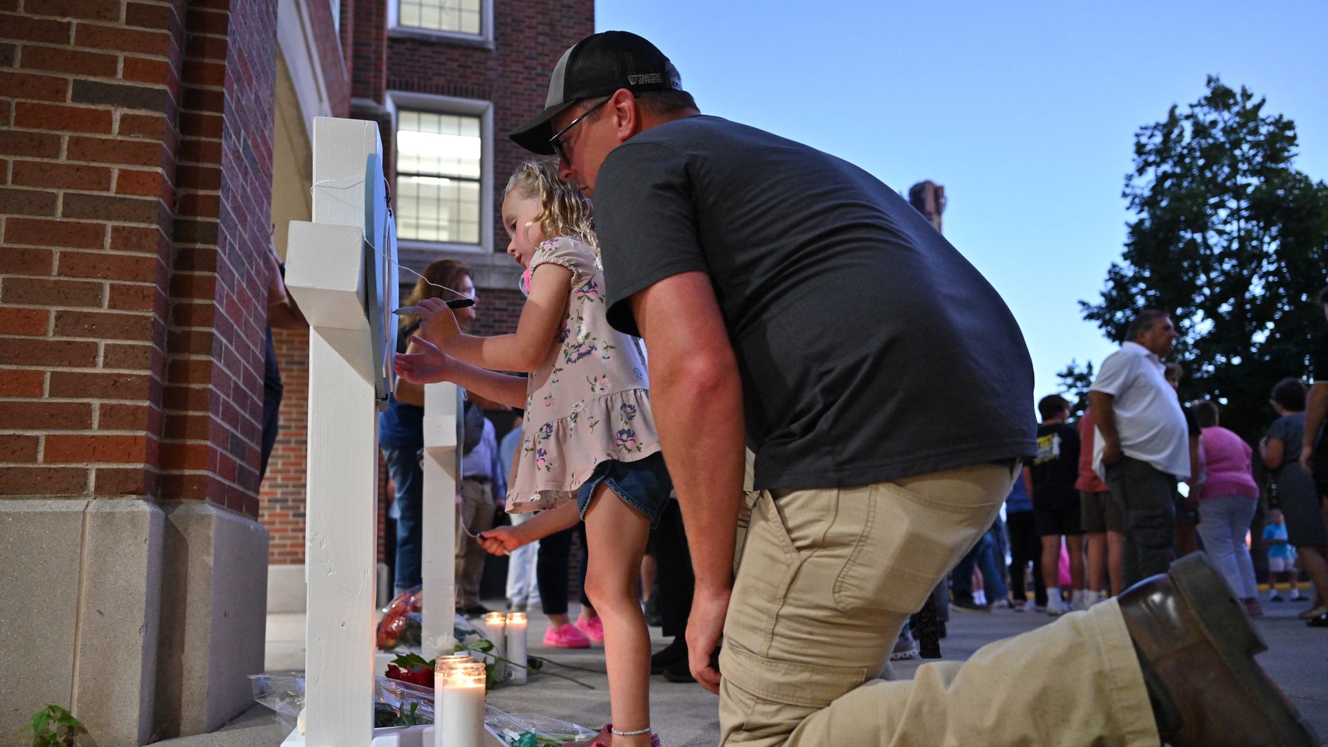 A man in a black shirt and beige pants kneels next to a young girl in a pink floral top and pink shoes writing on a white cross at a memorial with lit candles and flowers at dusk.