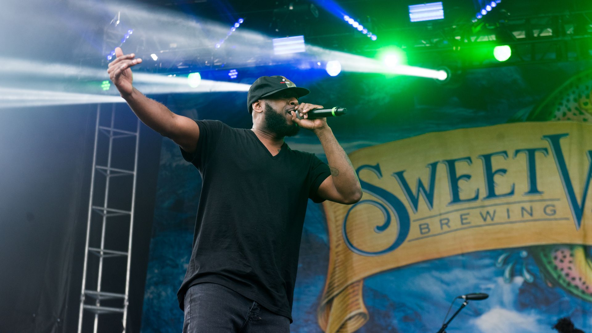 Man wearing black shirt and cap performing on stage with microphone, green and blue stage lights, and a backdrop displaying "SweetWater Brewing" logo.