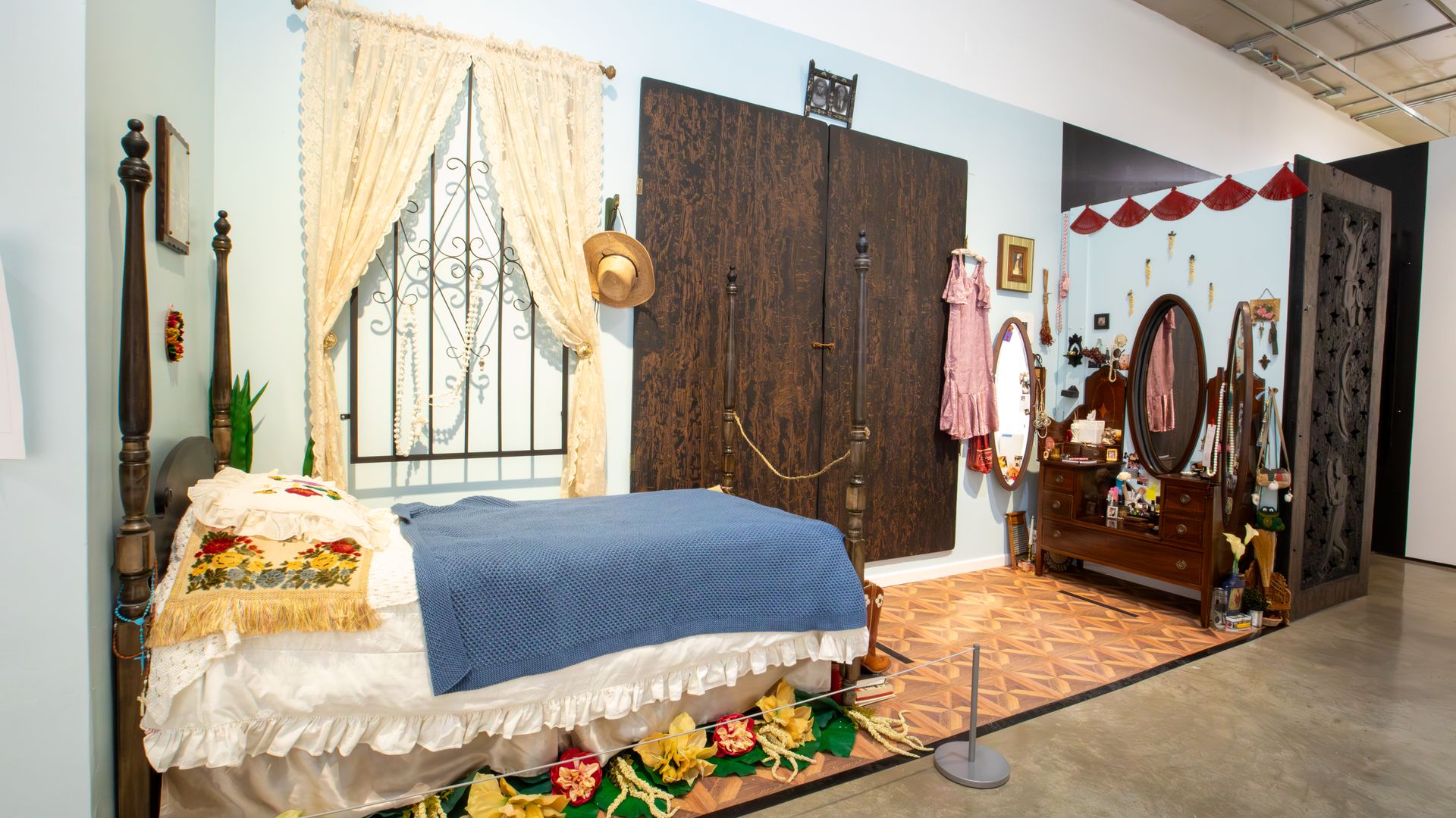 Vintage bedroom with four-poster dark wood bed, blue blanket, floral pillows, lace curtains, wooden dresser with three oval mirrors, pink dress hanging, and decorative flowers on floor.