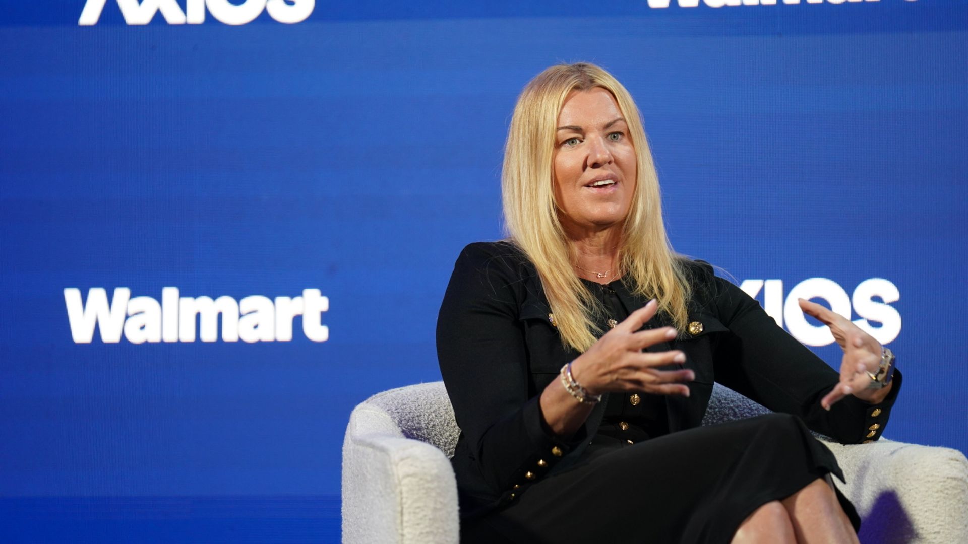 Blonde woman in a black outfit sits in a white chair on a blue stage with Axios and Walmart logos; she gestures with her hands while speaking.