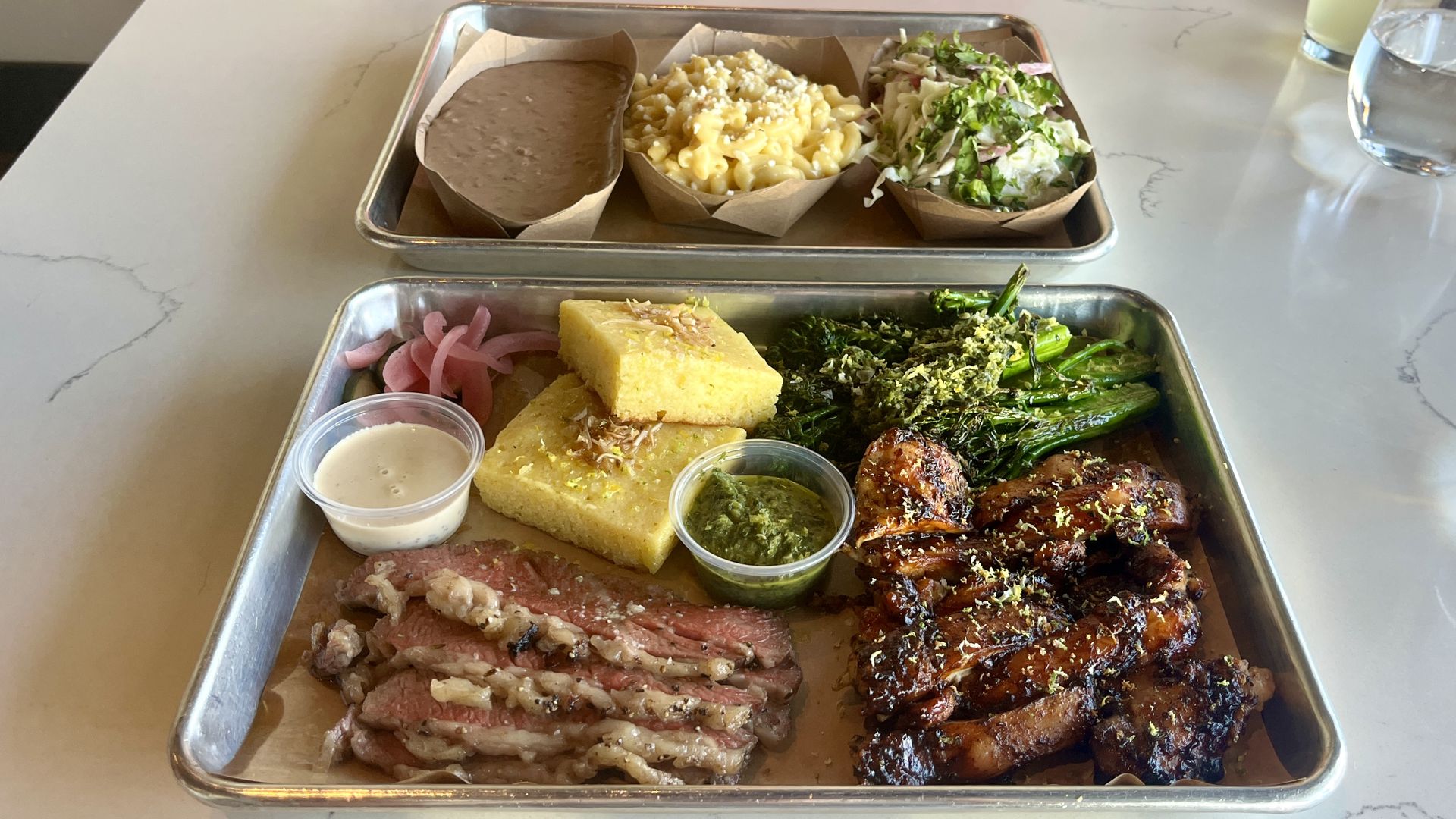 A tray of barbecue with smoked meats, broccolini, mac n' cheese, cornbread, pinto beans and slaw.