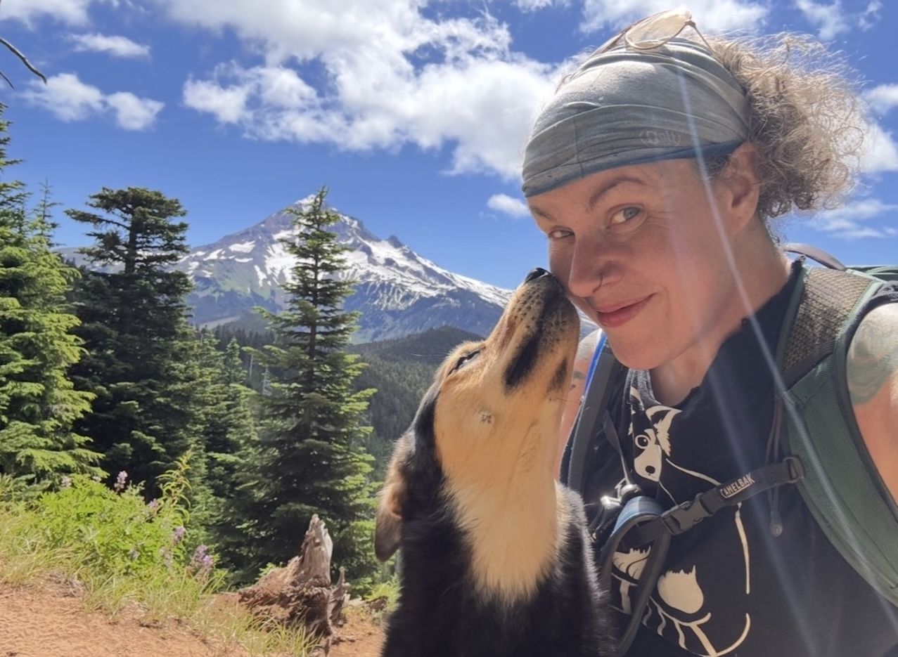 A person and a dog pose for a photo in front of a tree-filled forest peak, with a snow covered mountain in the background.