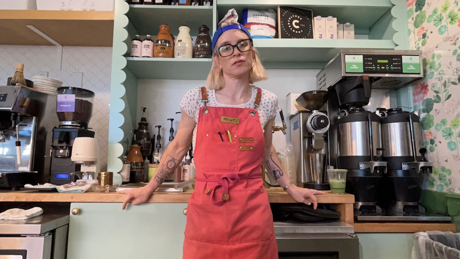 A woman stands behind a coffee bar with teal shelves, coffee machines, and floral wallpaper in the background.