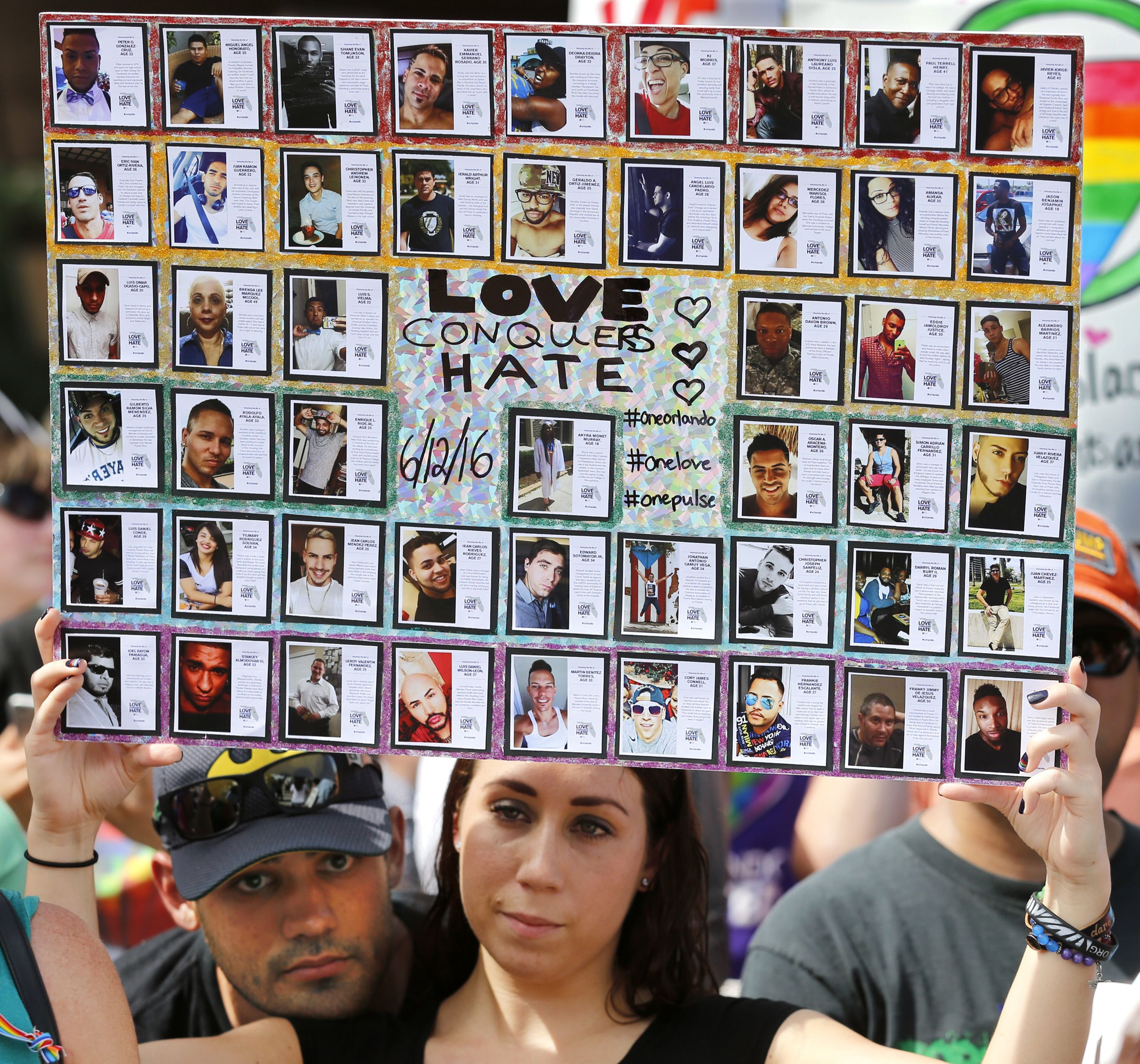 Colorful collage board covered with dozens of small framed photos of diverse people. Center reads "LOVE CONQUERS HATE" with hearts; two hands hold the board outdoors.
