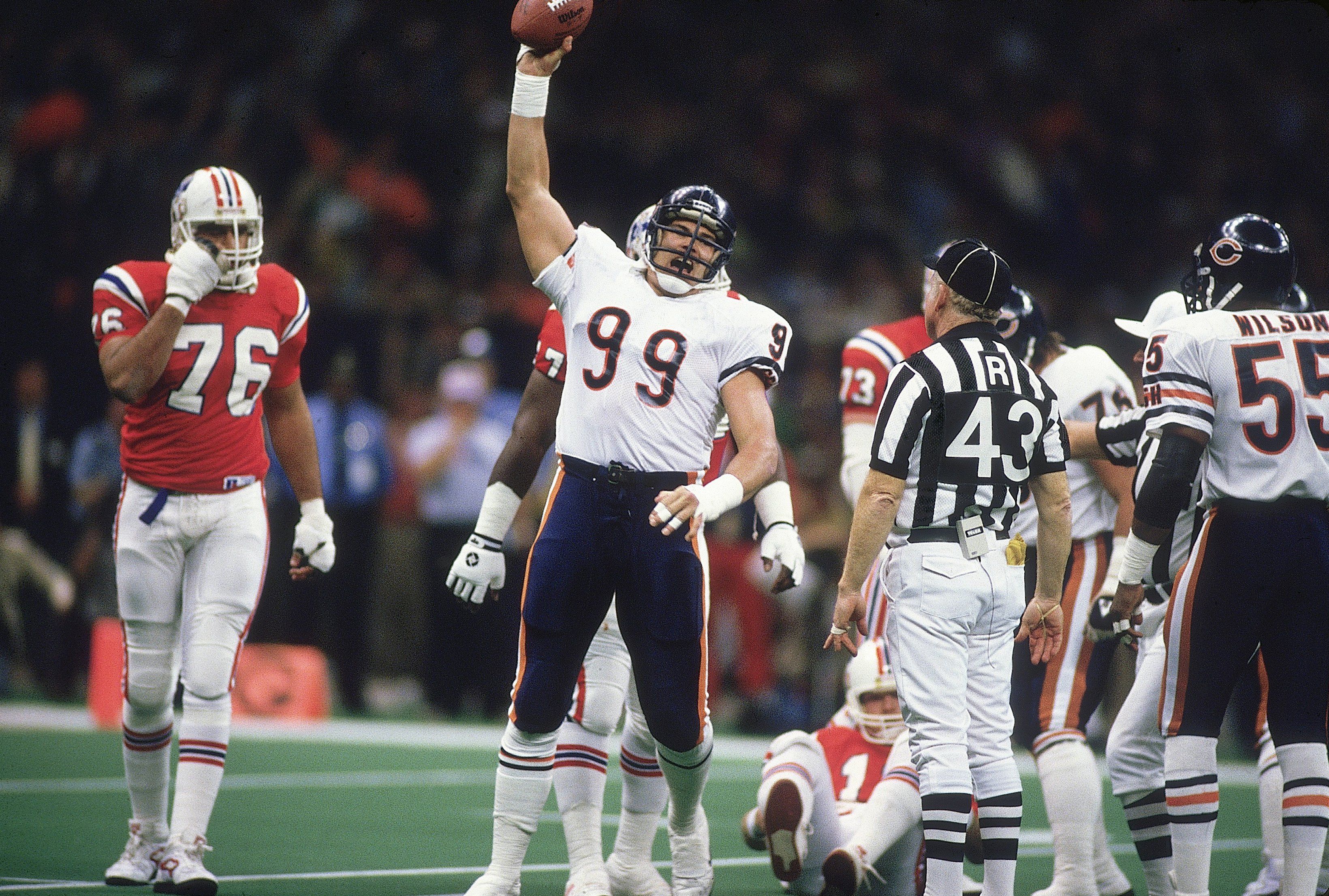 Chicago Bears player #99 raises football in celebration on field during game against New England Patriots, with referee #43 and other players in red and white uniforms nearby.