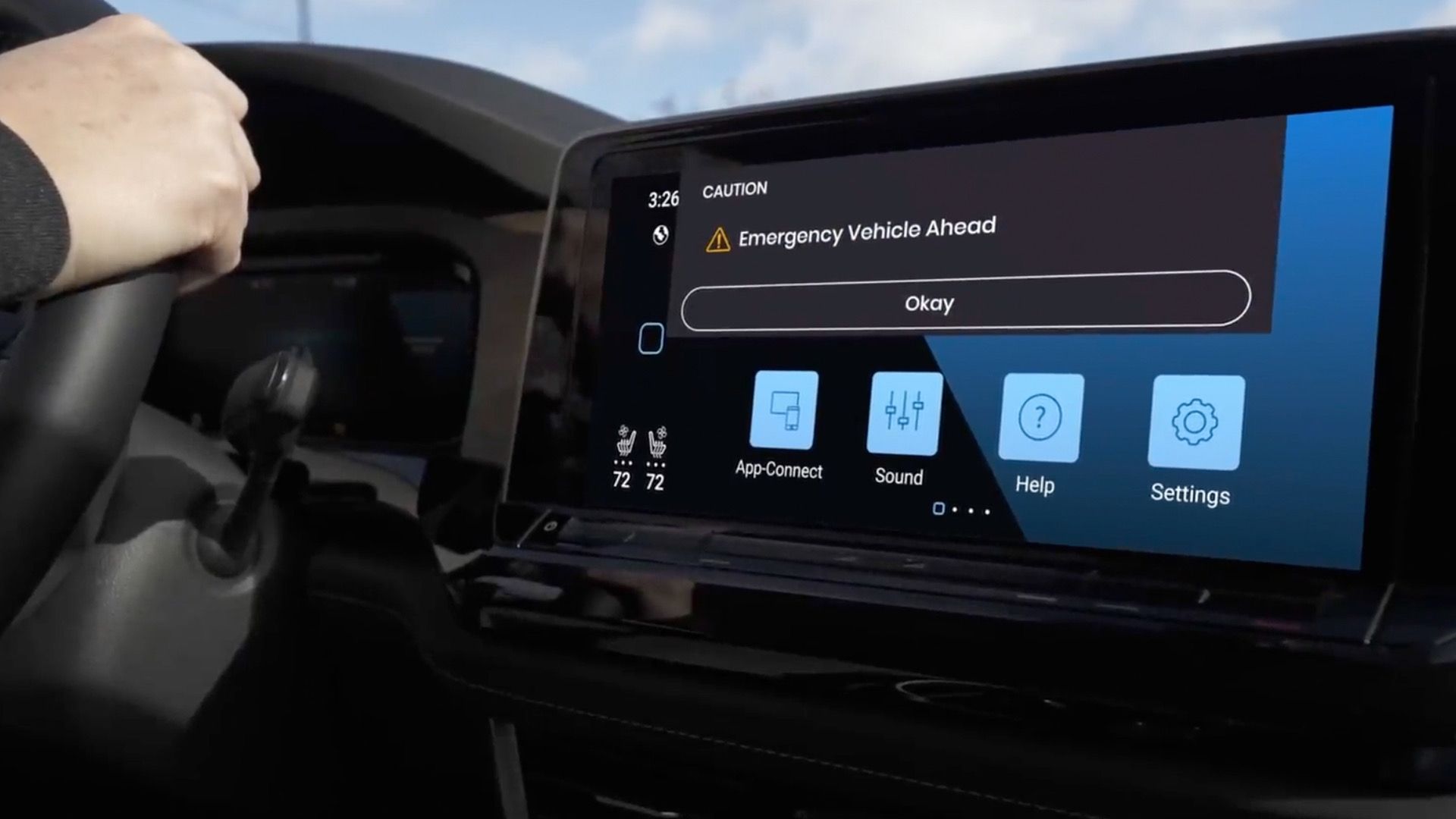 Driver’s hand on the steering wheel as the car’s touchscreen shows a CAUTION: Emergency Vehicle Ahead alert with an Okay button, plus bottom icons for App-Connect, Sound, Help, and Settings.