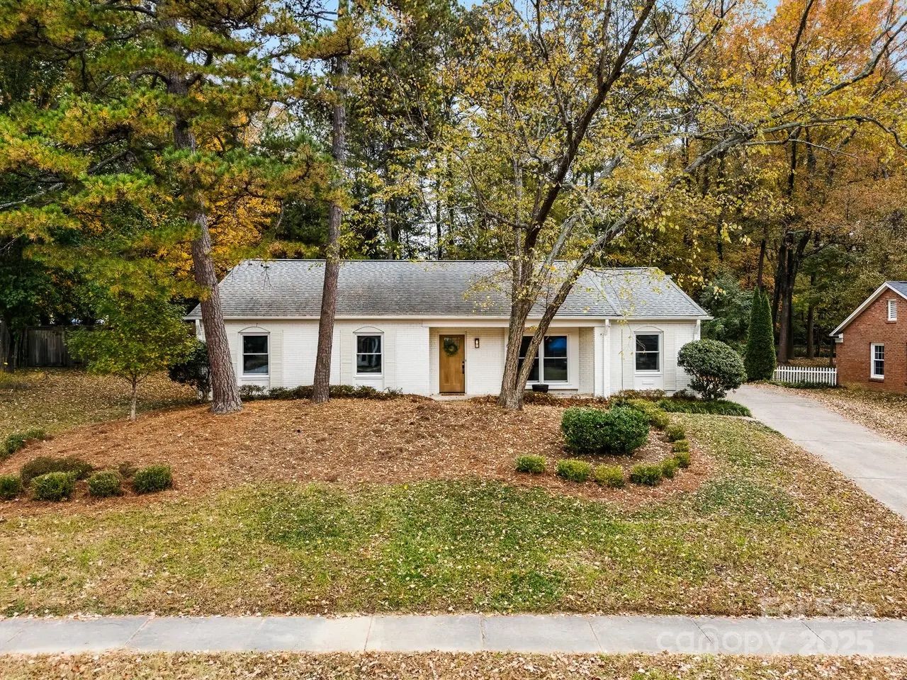 Single-story white brick house with a gray roof, surrounded by trees with autumn foliage and a front yard featuring mulch and small green bushes along a concrete driveway.