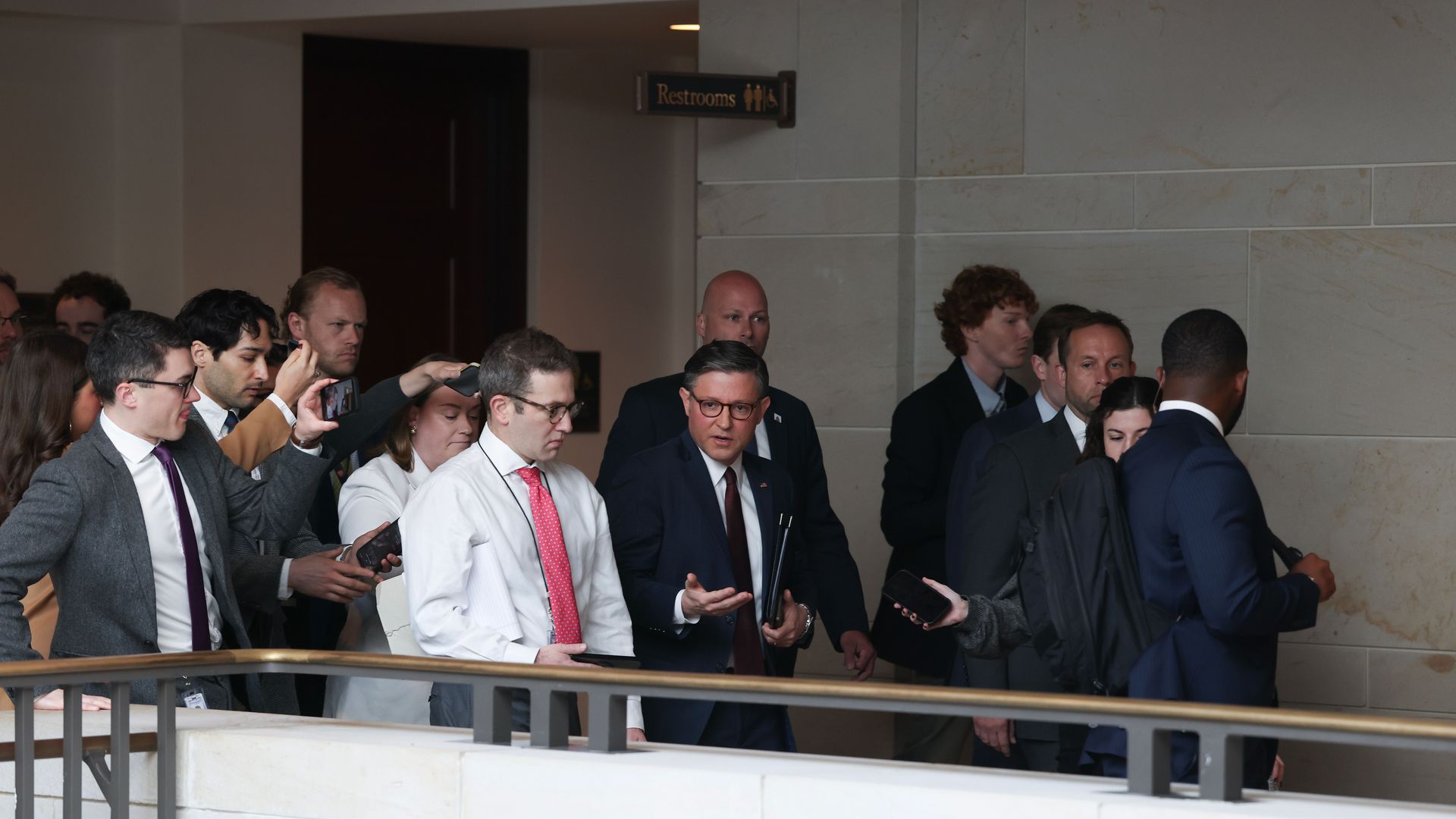 Group of professionally dressed people walk along a marble hallway. A man in a navy suit with a red tie gestures while holding folders; others film with phones, near a railing and a restroom sign.