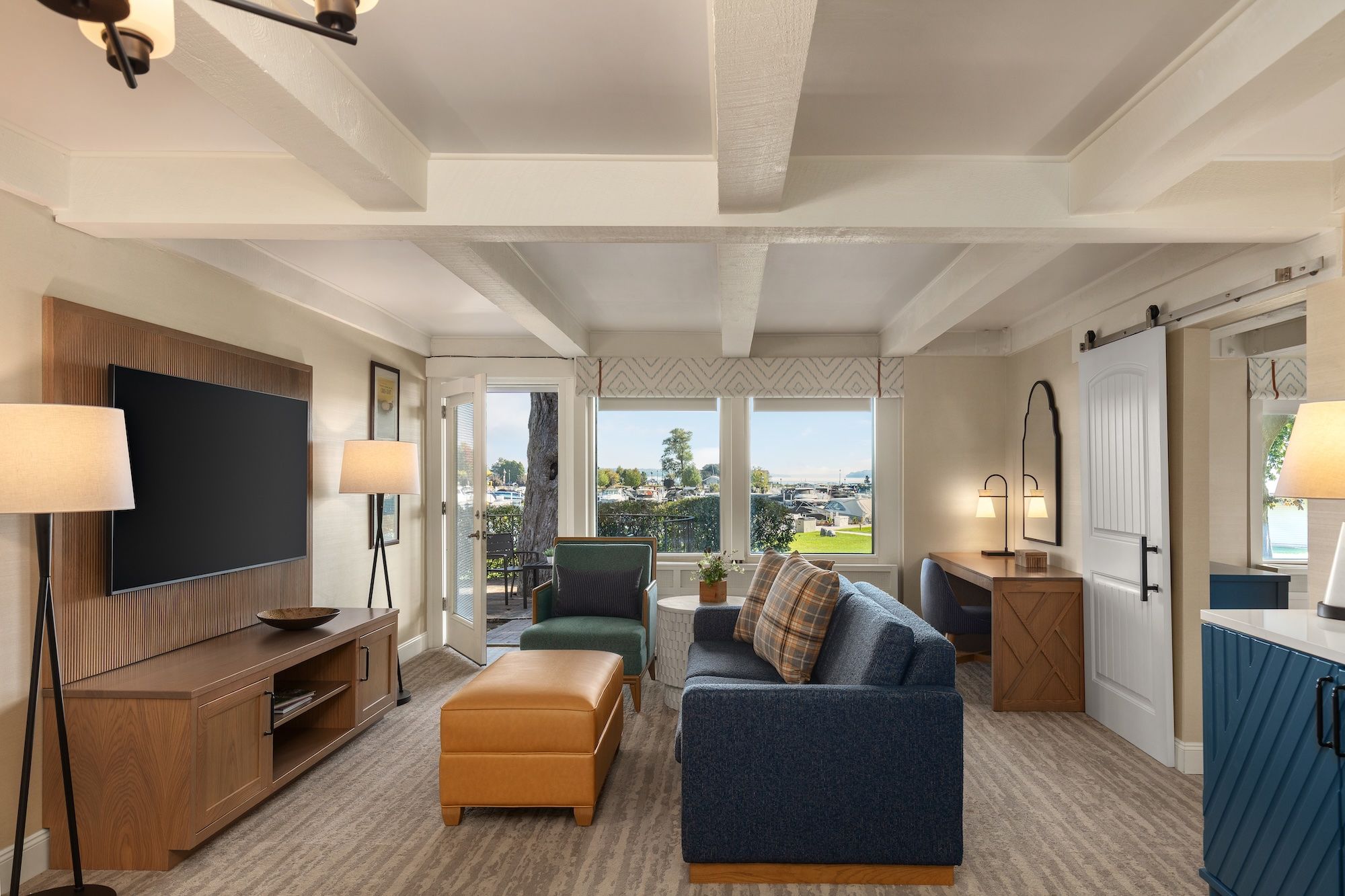 Living room with white exposed ceiling beams and beige walls; large windows overlook a waterfront with boats. Blue sofa, green chairs, orange ottomans, and a wooden media console.