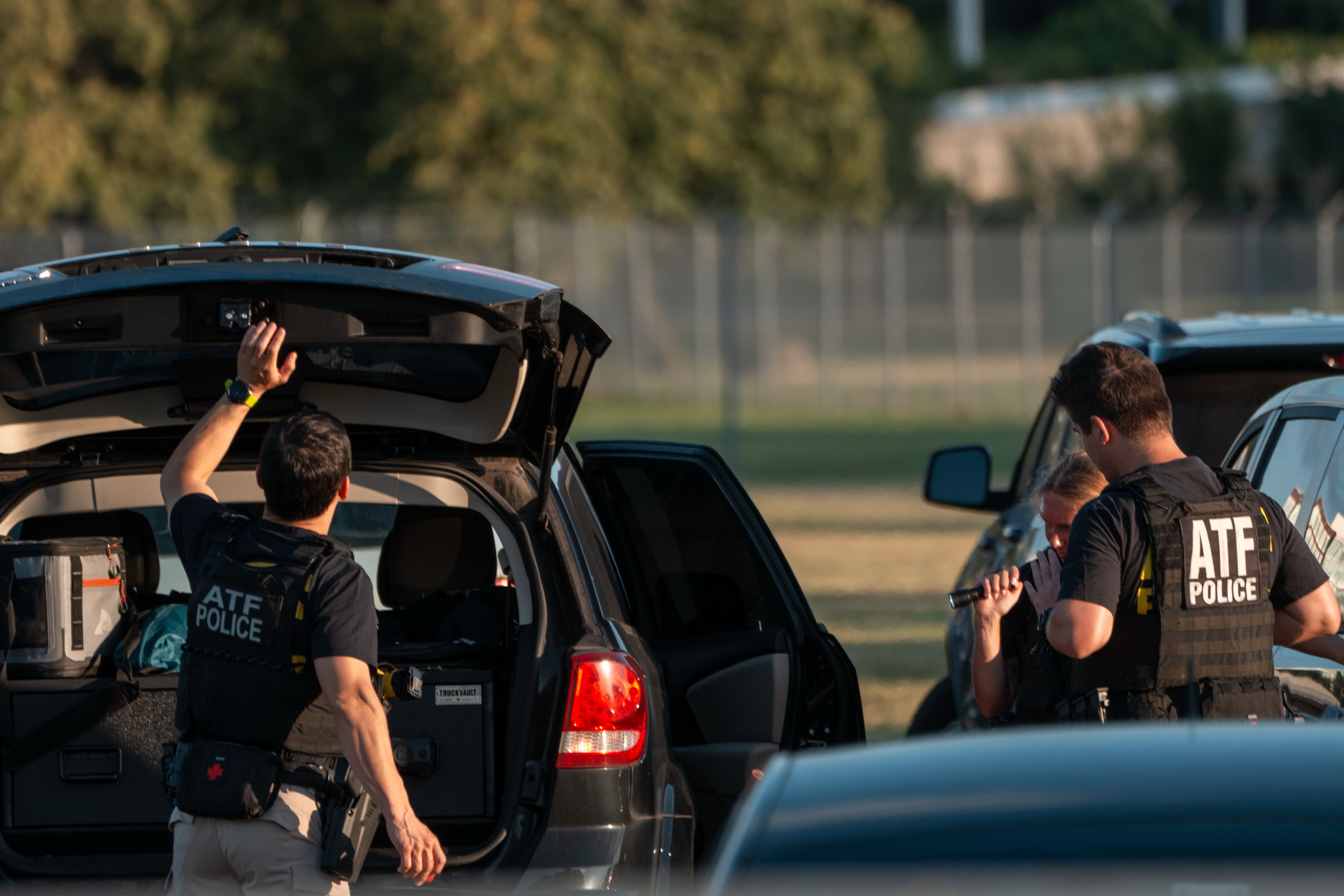 Two ATF police officers in black tactical vests stand near black SUVs with open doors and trunk, one officer holding the trunk open in an outdoor, grassy area with blurred trees and fence background.