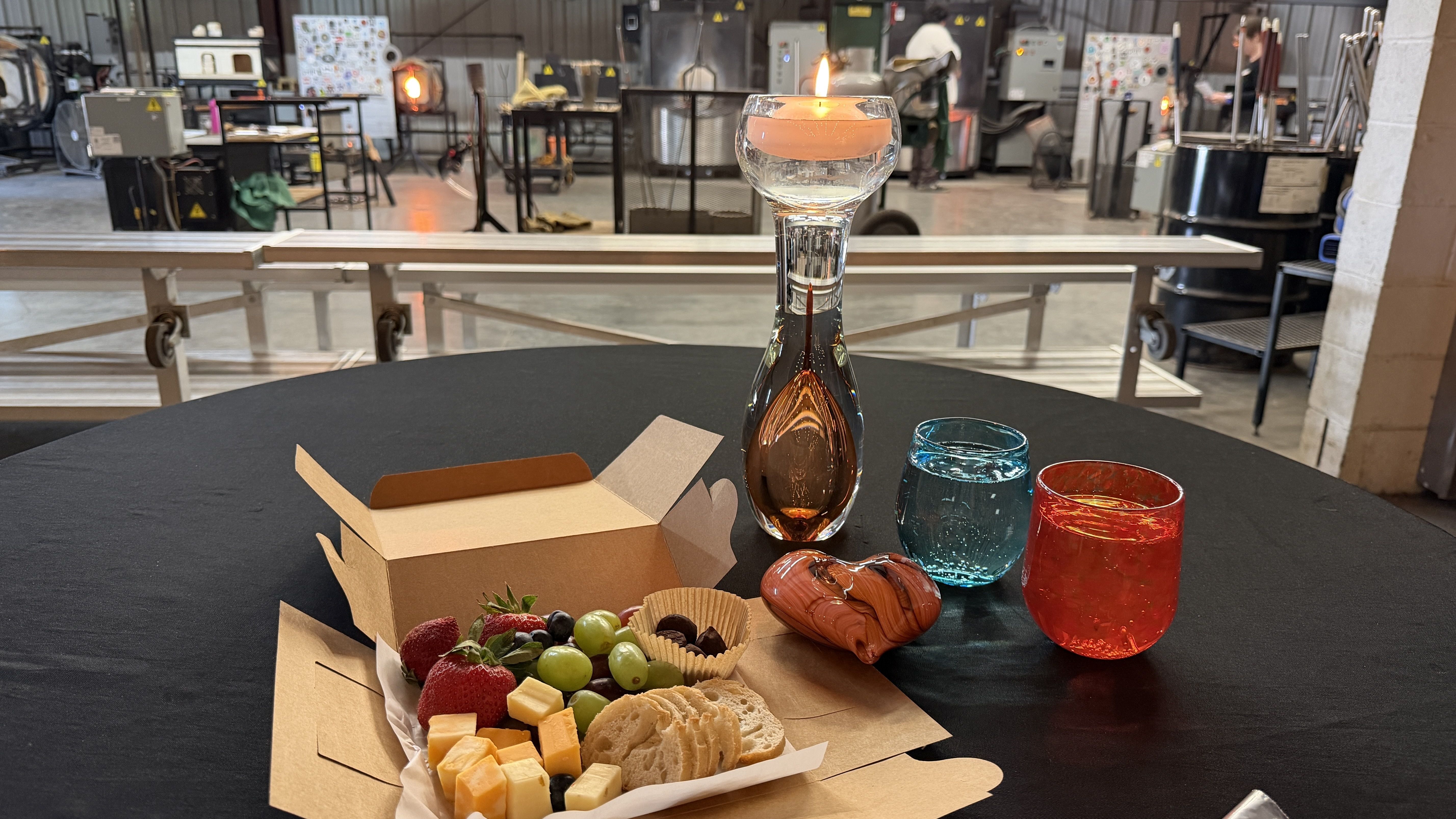 Round black table with an open cardboard box of fruit, cheese, and crackers. A tall glass decanter with amber liquid and a lit candle on top. Blue and red glasses sit nearby; industrial workshop background.