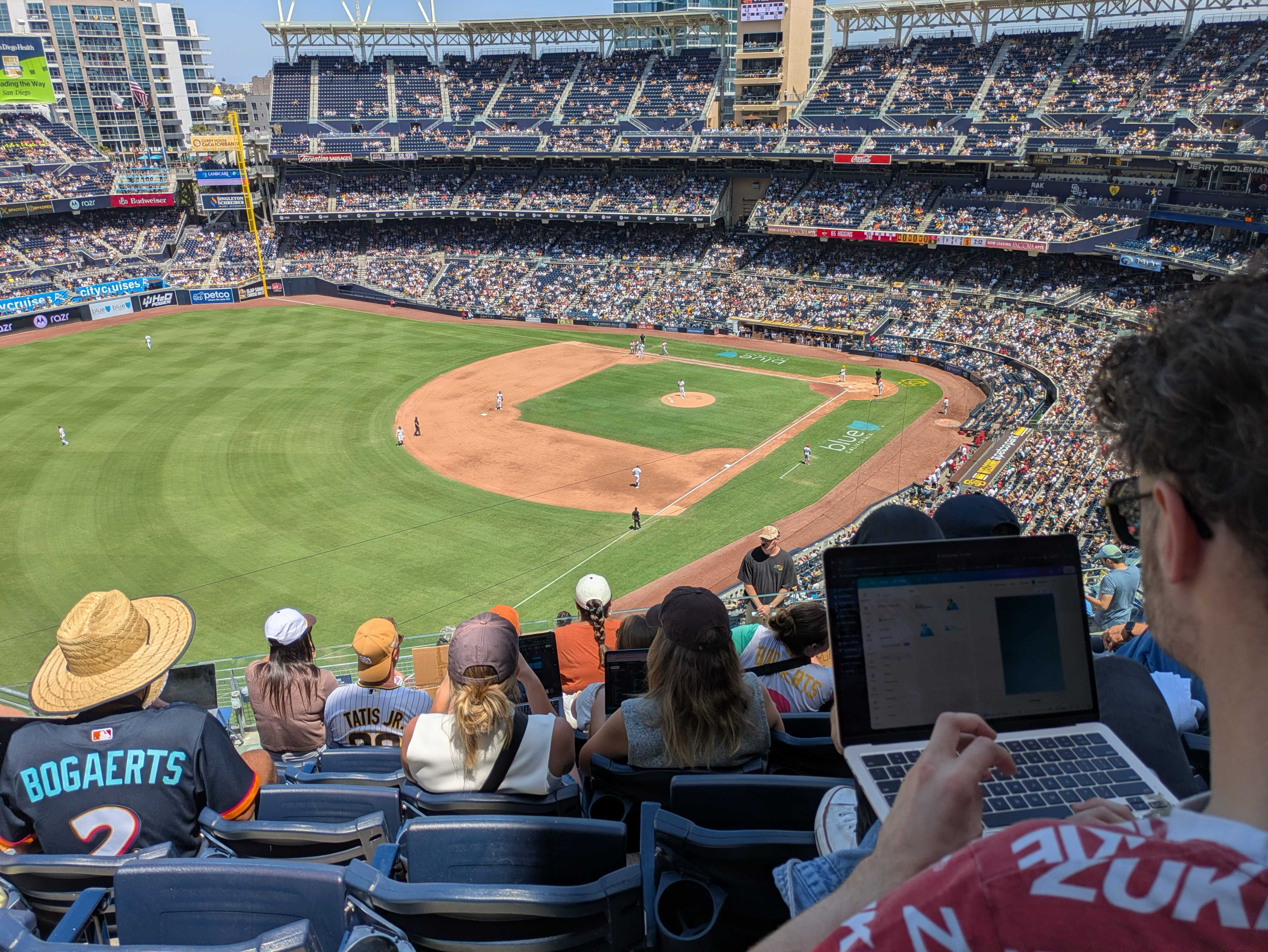 Wide view of a sunny baseball stadium packed with fans in blue seats; players on the brown infield near home plate; foreground fans wear hats and jerseys, some with laptops.