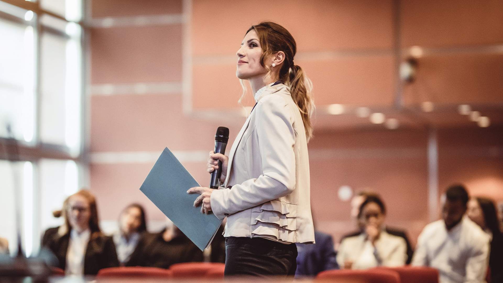 A woman carrying a folder and microphone, speaking in auditorium. 