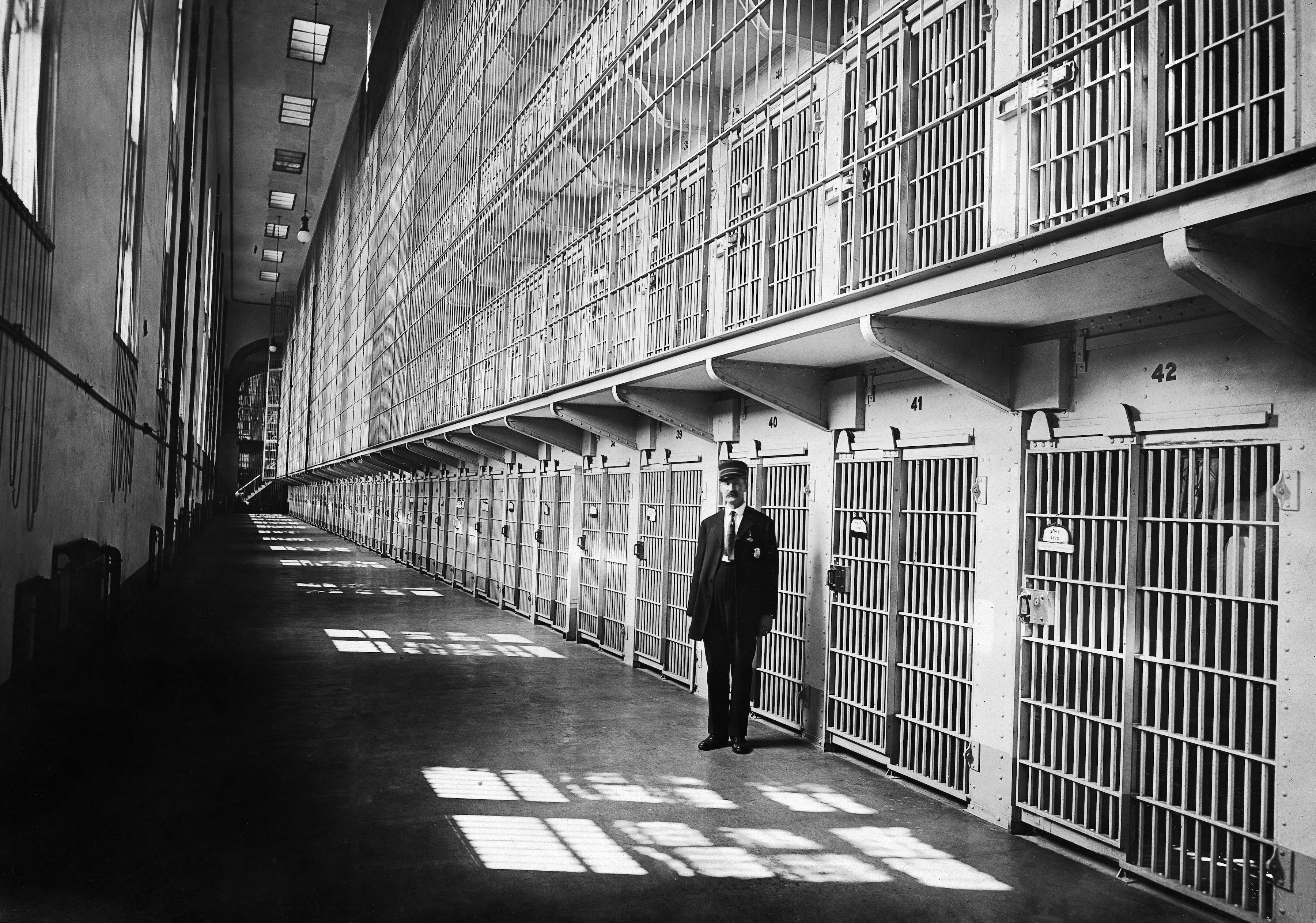 A black-and-white image of a long prison corridor with barred cell doors along both walls. A suited guard stands near the foreground as overhead lights cast striped shadows on the floor.