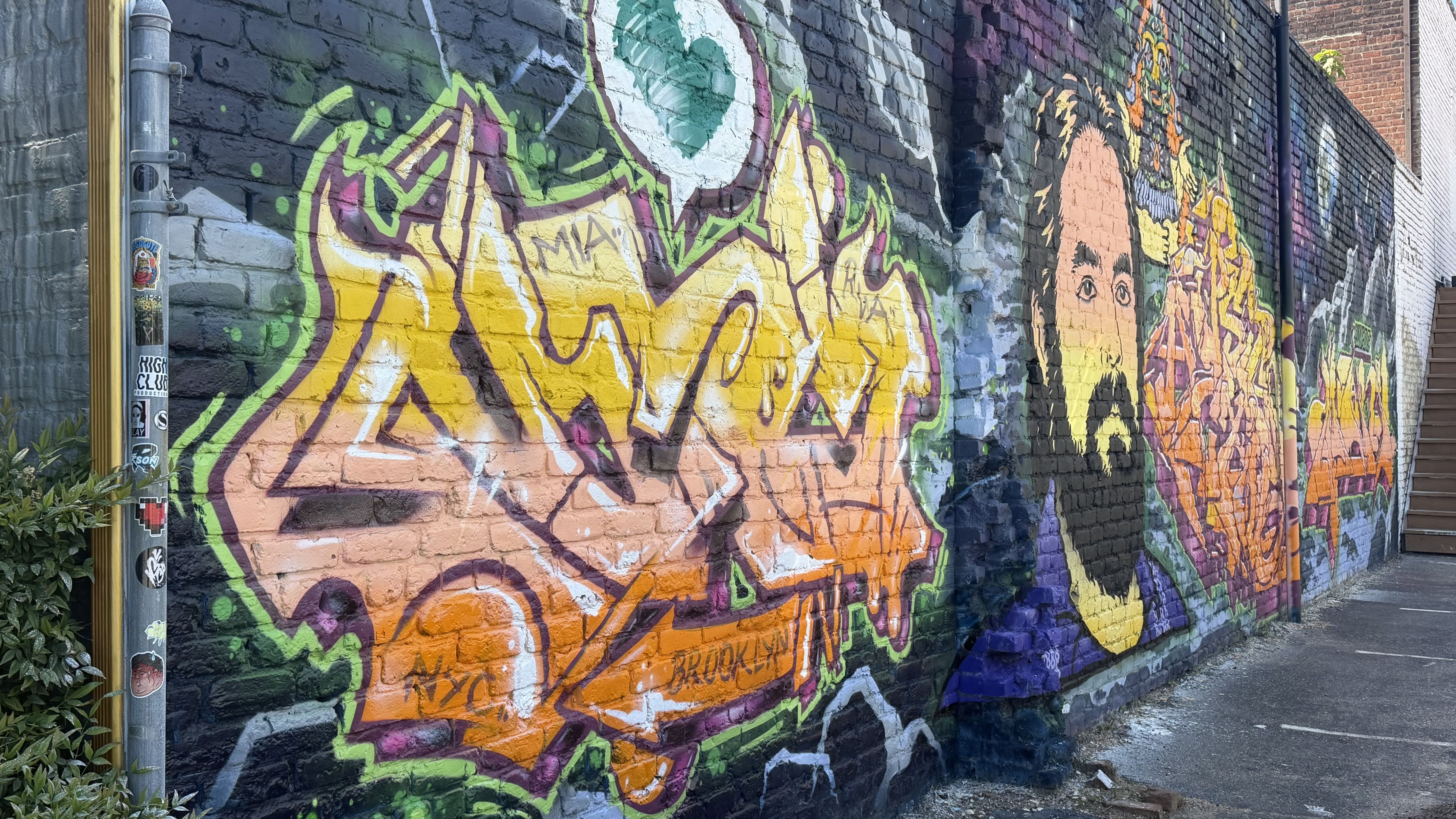 Wide alley wall covered in bright graffiti on brick, with yellow, orange, and green lettering and cartoon faces; a stickered utility pole on the left; stairs on the right; wet ground.