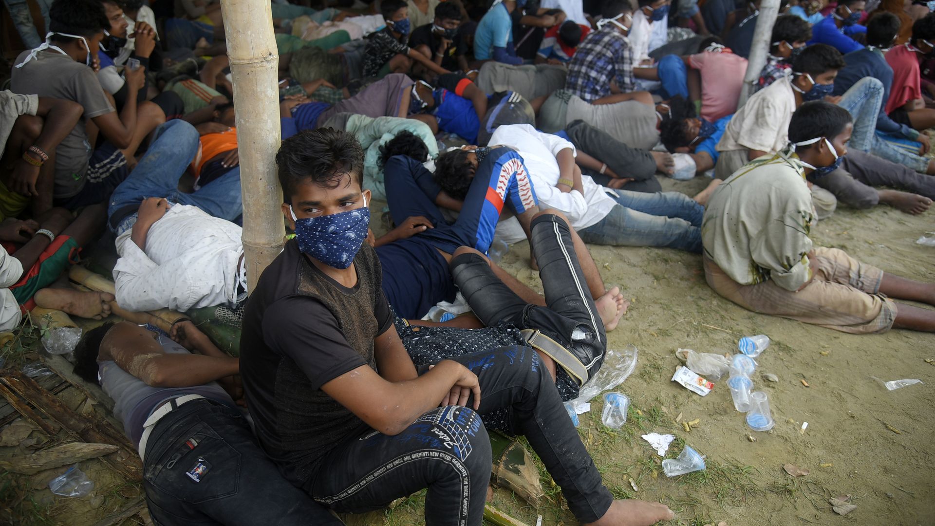 A boy wearing a face mask sits in the dirt in front of a crowd of people laying on the ground or sitting
