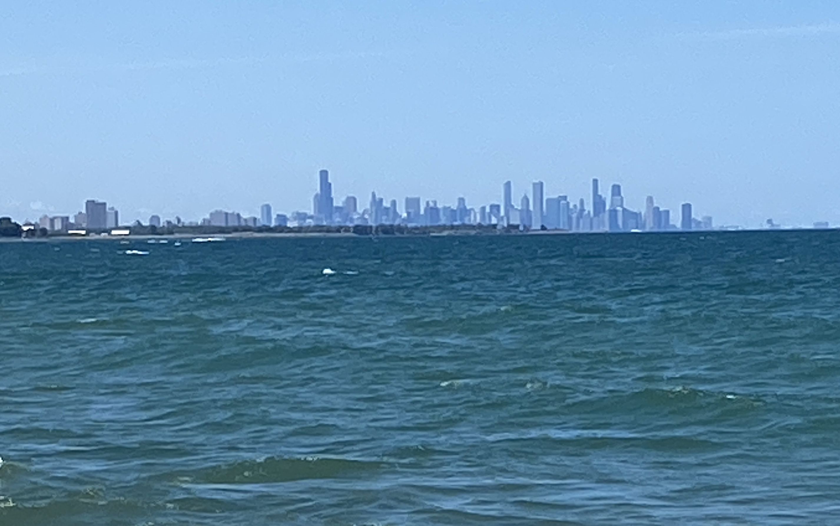 Distant city skyline across a blue sea, tall rectangular skyscrapers silhouetted on the horizon under a clear sky. Dark green water with small whitecaps in the foreground.