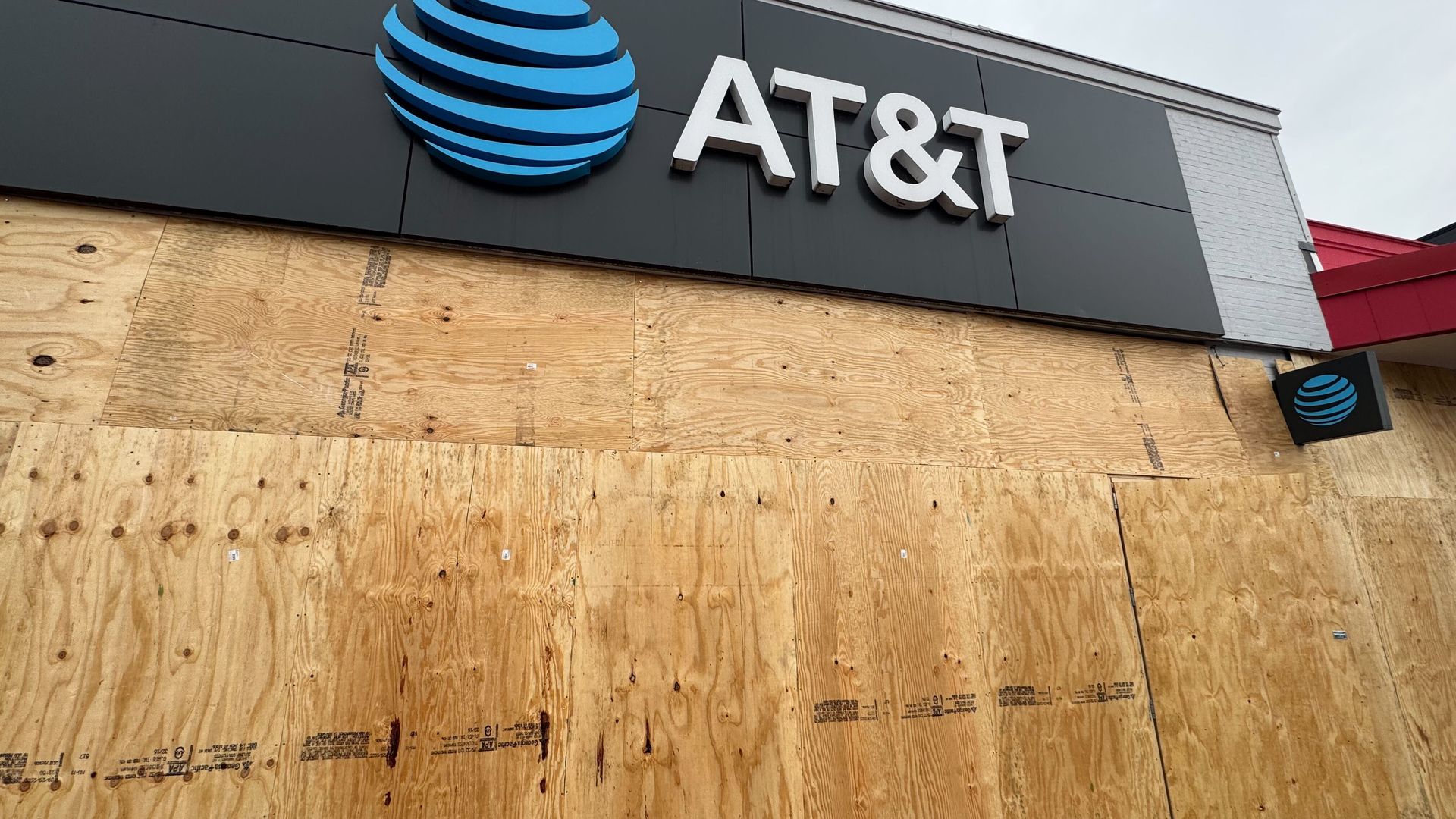 AT&T store with large blue and white logo above entrance, windows and door covered with plywood boards, cloudy sky overhead, concrete sidewalk in front.