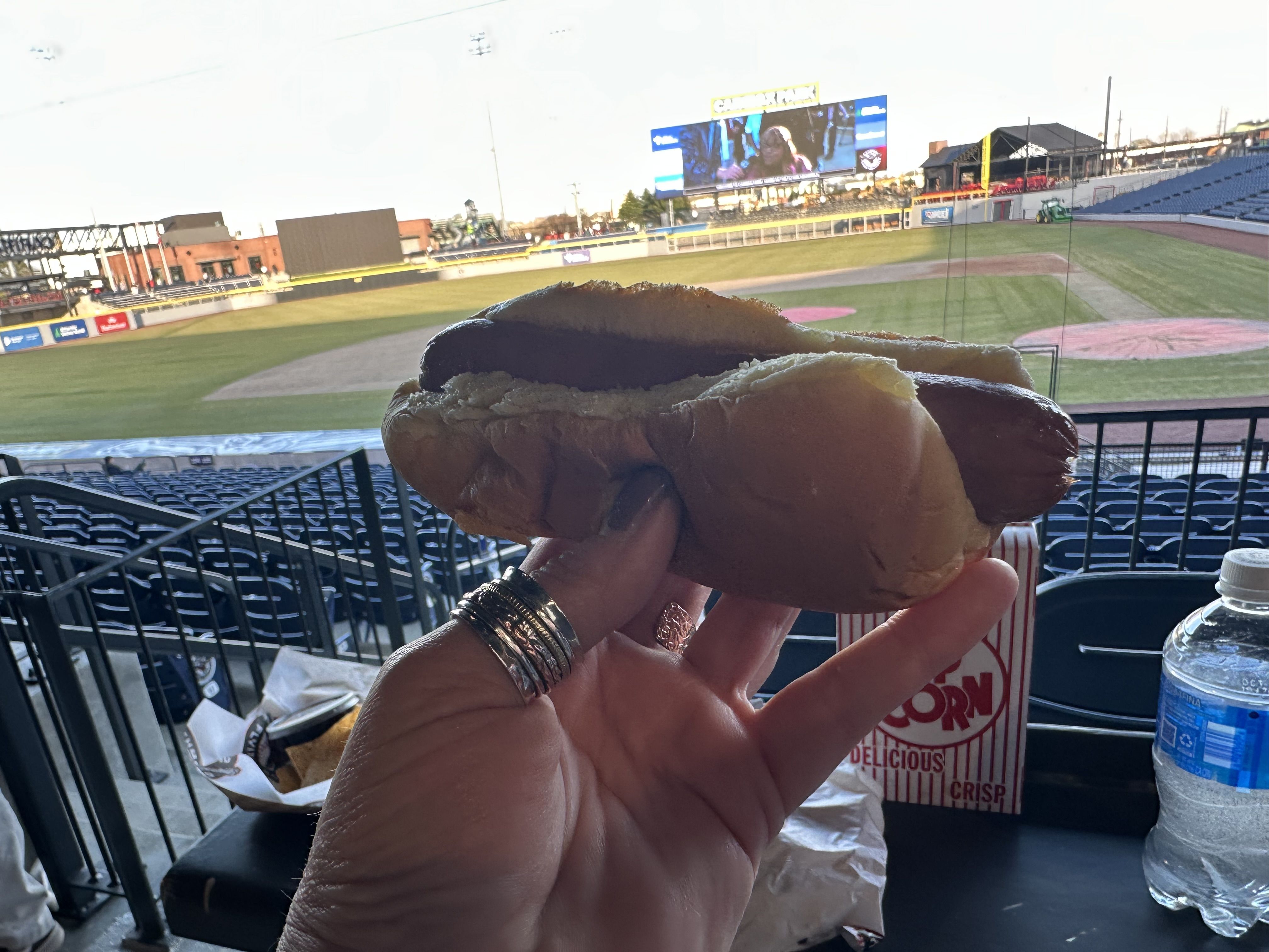 Hand with silver rings holds a giant sandwich in a baseball stadium view: green field, blue seats, a big video screen, and a red-and-white popcorn cup nearby.
