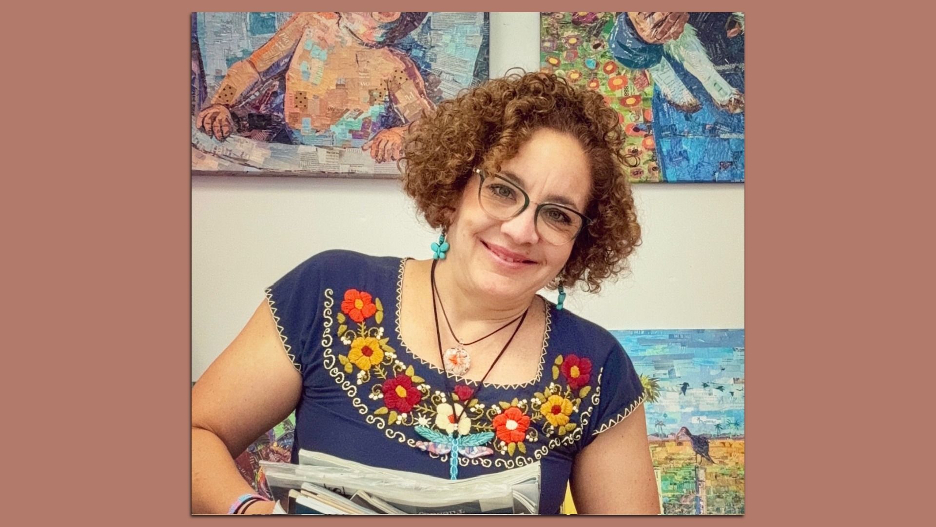 Woman with curly hair, glasses, and turquoise earrings smiling. She wears a navy blue shirt with colorful embroidered flowers and holds a clear bag with various items inside.