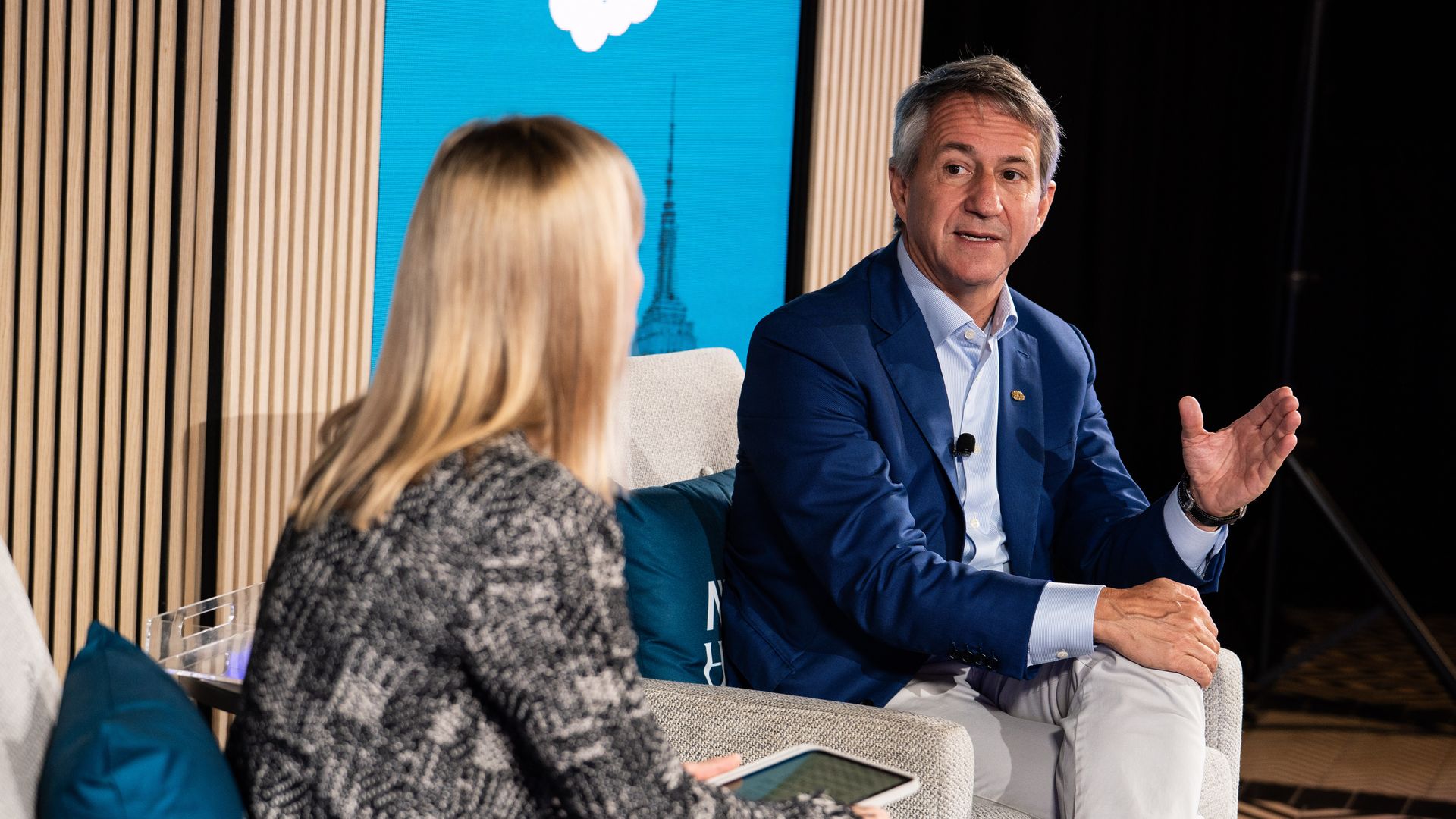 A man in a blue blazer gestures while speaking to a woman holding a tablet, seated on chairs with a blue "salesforce" logo screen behind them in a modern setting.