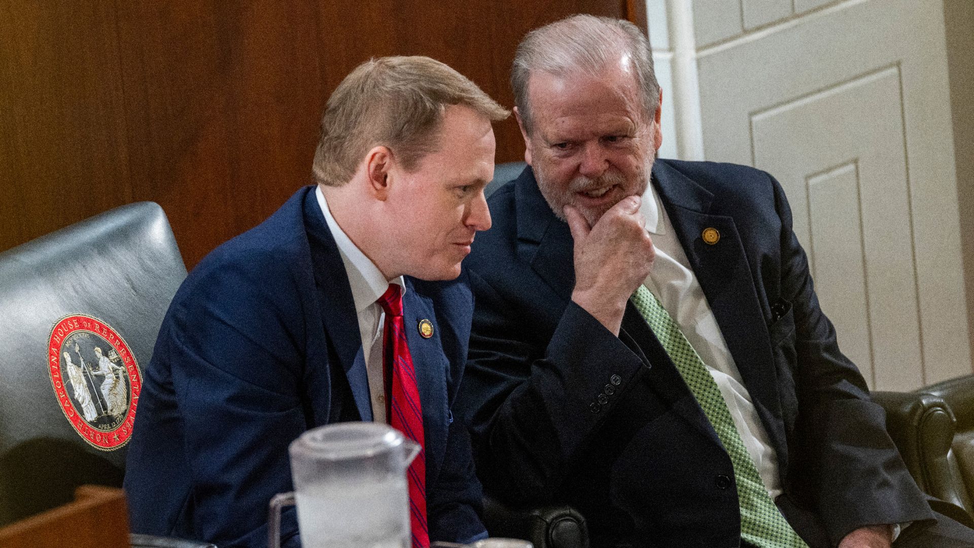 Two men in suits sit closely together, leaning in for a private conversation during a formal government session. One man, wearing a red tie, listens intently while the other, with a green tie, smiles and rests his hand on his chin.