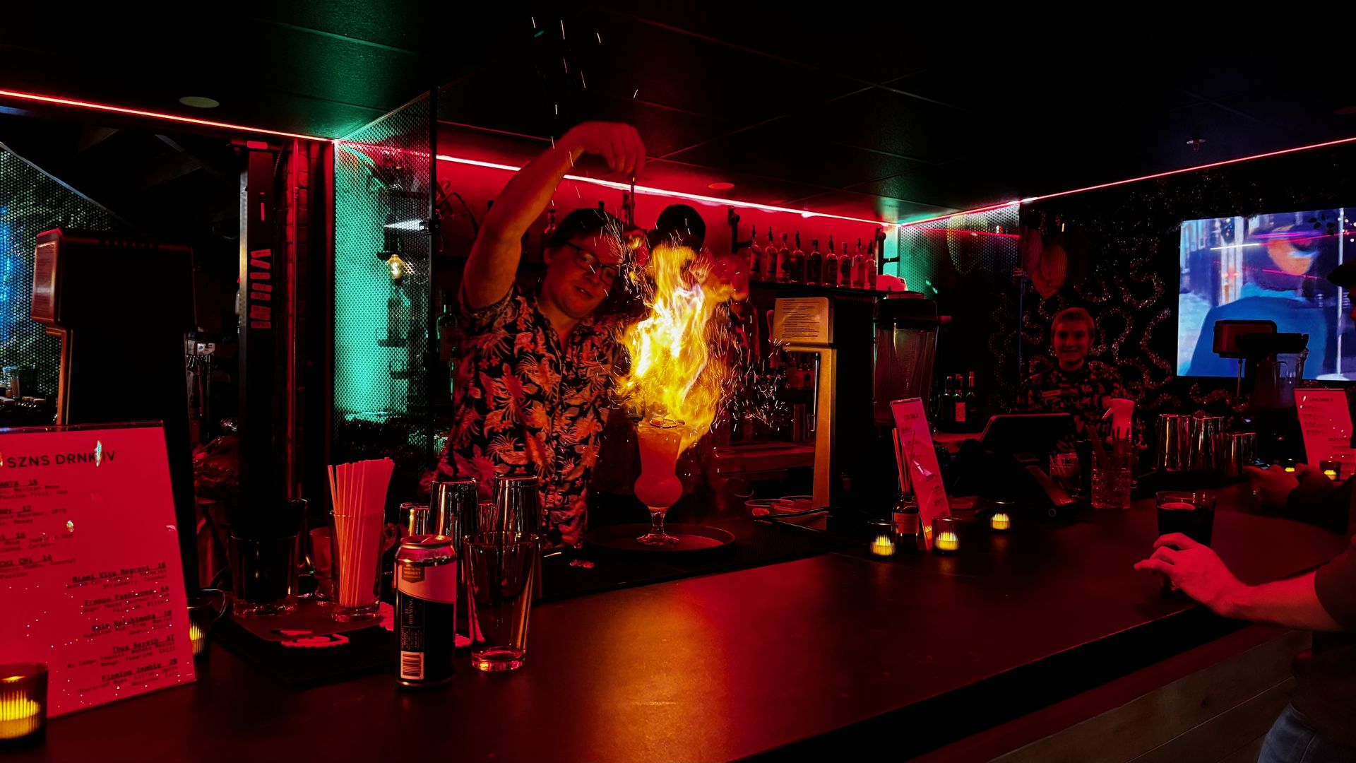 Bartender in a floral shirt creates a flaming cocktail at a dimly lit bar with red and green neon lights, while a smiling bartender and a person with a drink are visible in the background.