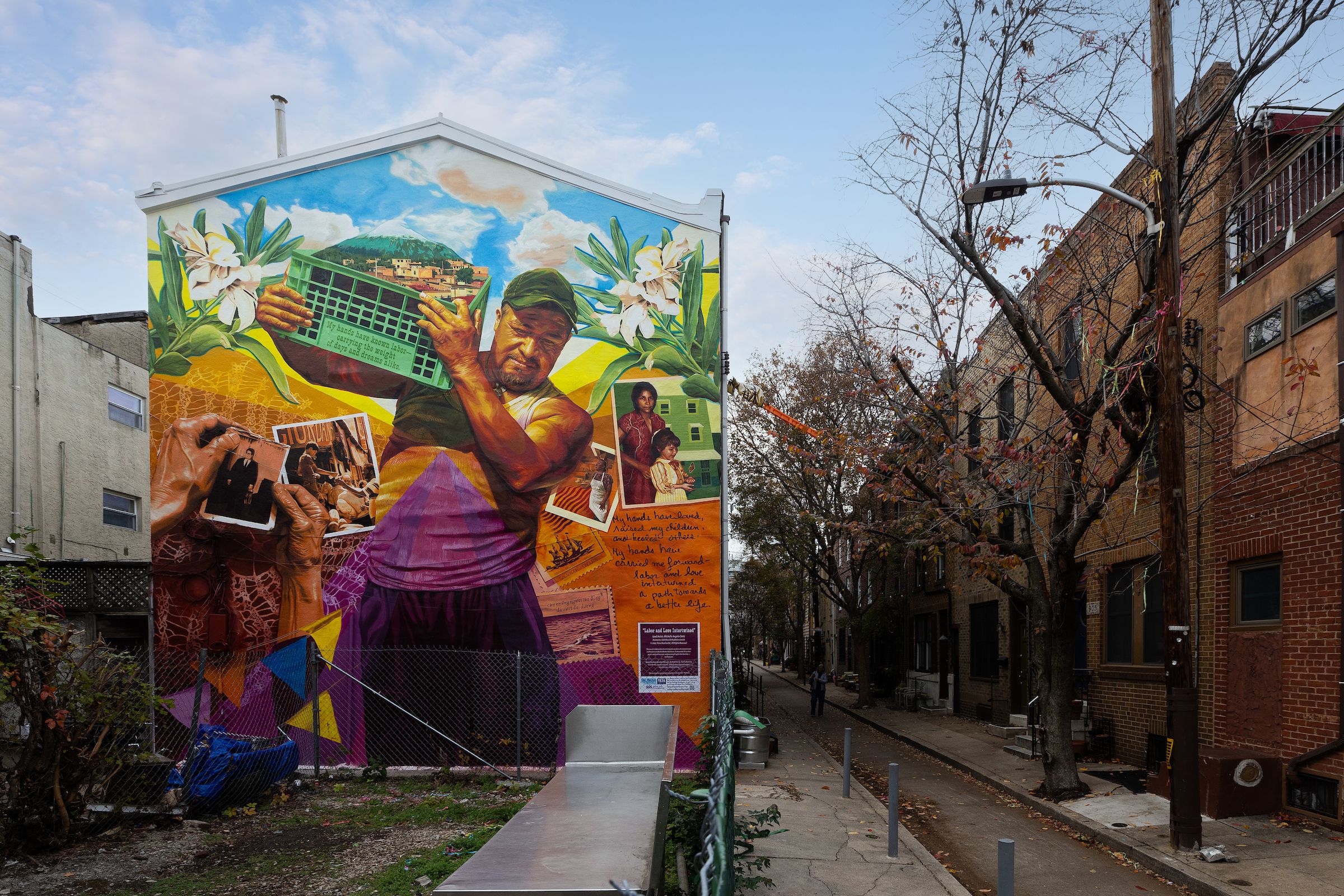 A mural of a farm worker holding a basket with a mountain and village in it, along side photos of people and under a blue sky with puffy clouds. 