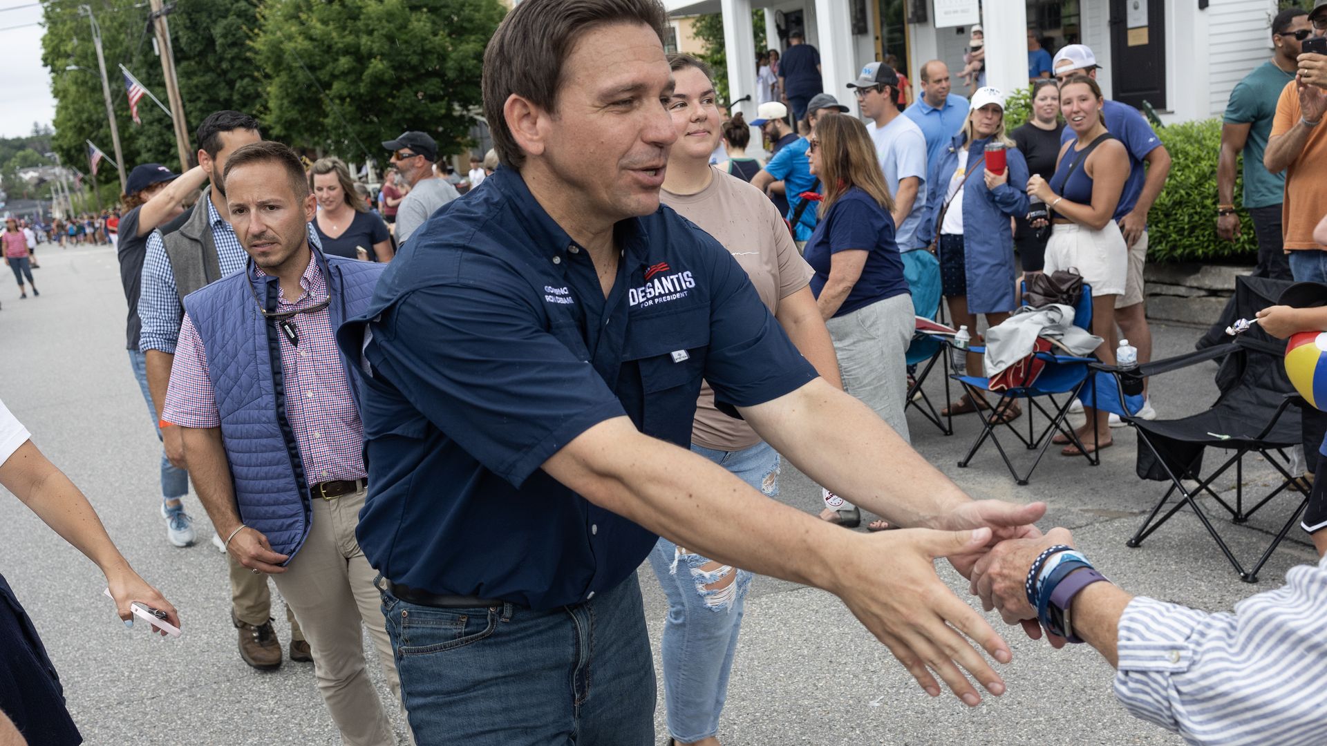 A man in a navy blue shirt that says "DeSantis for president," surrounded by people, shakes hands with someone out of frame.