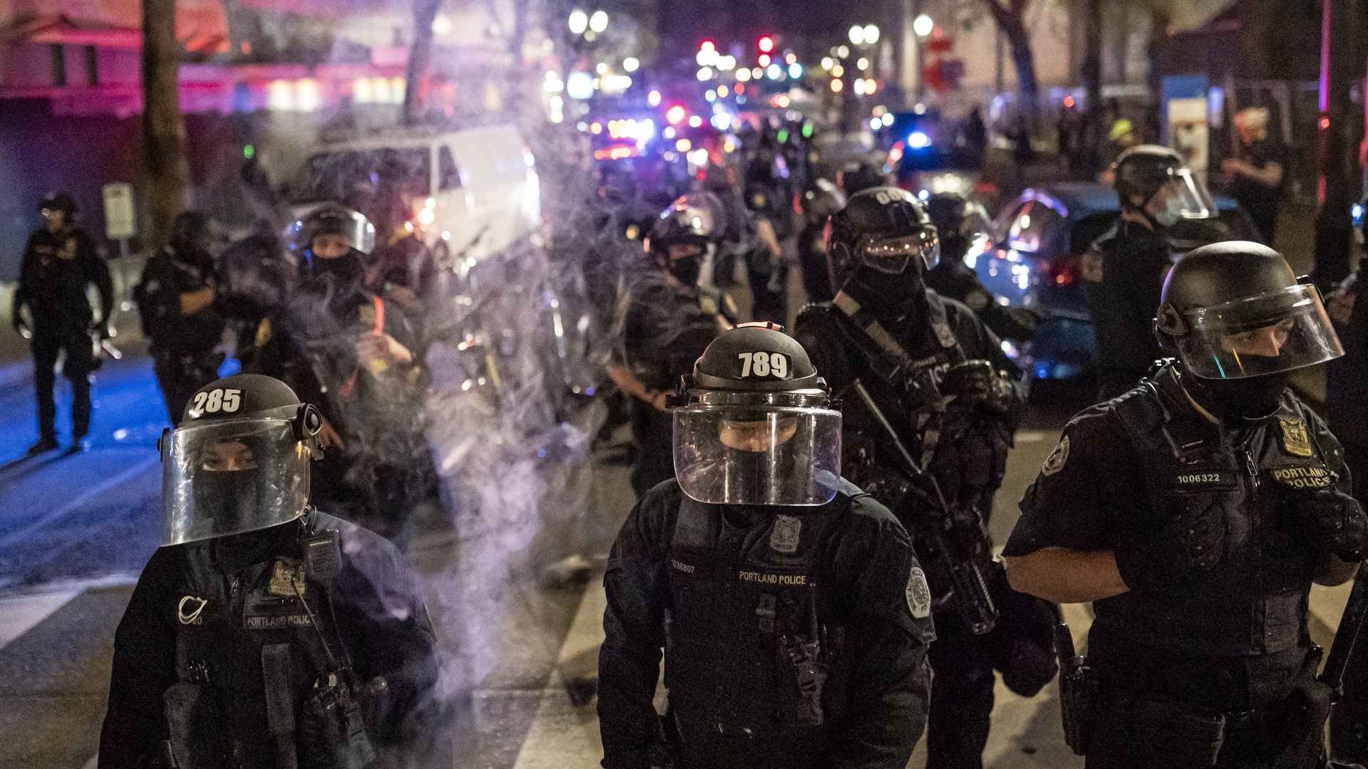 A line of police dressed in riot gear walk down a street with tear gas hanging in the air 