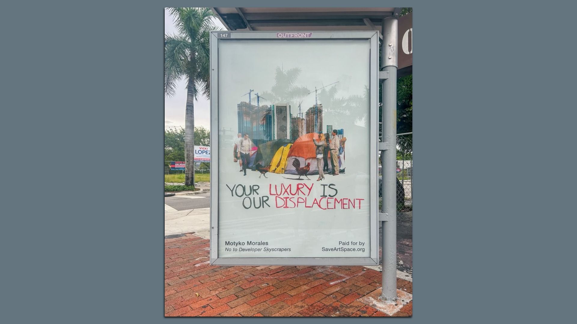 Bus stop poster shows a city skyline with cranes, tents, and people; the caption reads "YOUR LUXURY IS OUR DISPLACEMENT" in black and red, with palm trees in the background.