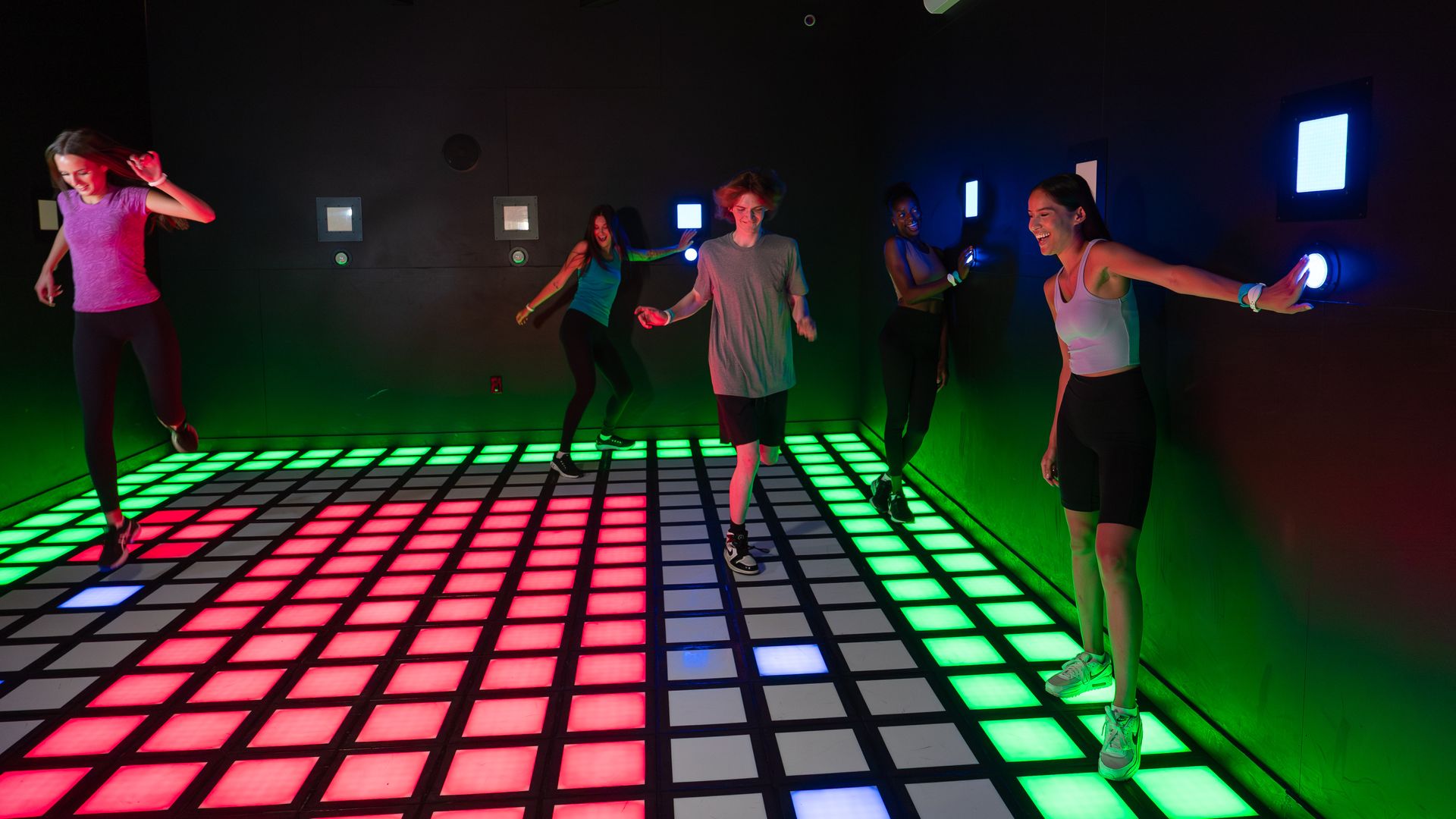 Several people jumping on a large grid composed of lighted electronic squares of various colors in a a darkened room. 